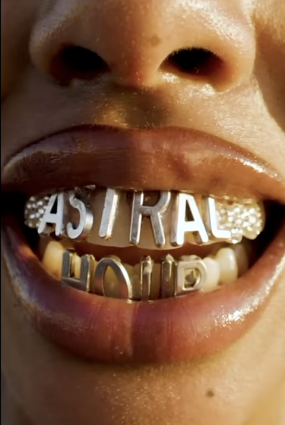 Close-up of a person's mouth showing gold teeth spelling out 'FAME' with tongue and lips slightly open.