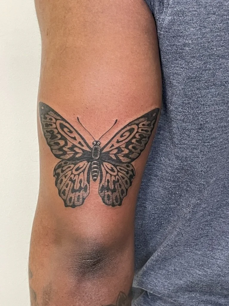 Tattoo of a butterfly on person's upper arm, with detailed black and gray wings and body.