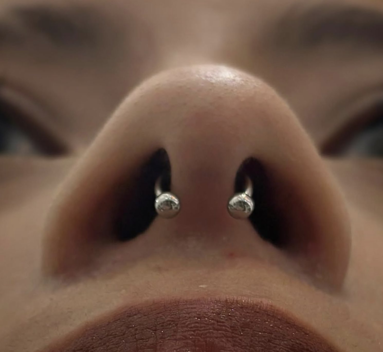 Close-up of a person's nose with two silver nose rings and a person's closed lips visible at the bottom of the image.