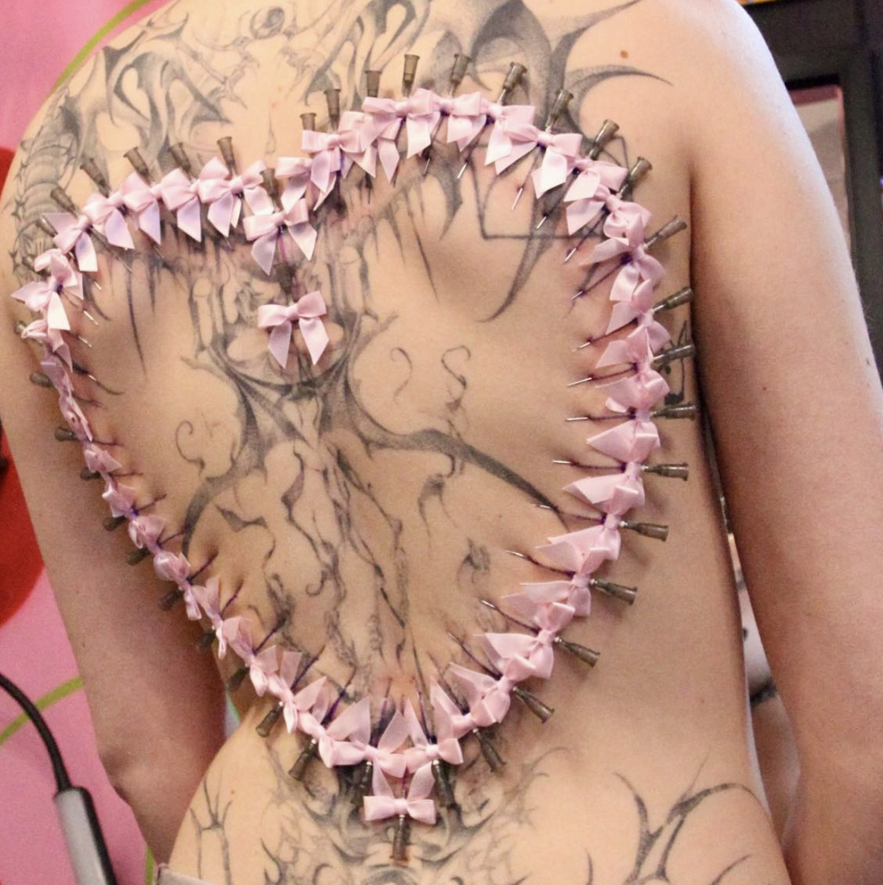 A tattoo of a tree on a person's chest with pink bows and metal pins around the outline of the tree.