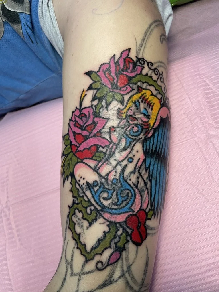 Tattoo of a pin-up girl with blonde hair, blue eyes, and pink lipstick, surrounded by roses and decorative swirls, on a person's arm.