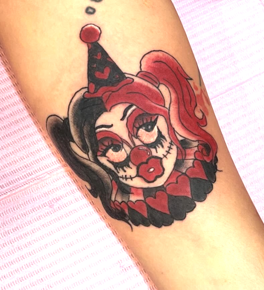 Tattoo of a female clown with red hair, black hair, and makeup, wearing a hat with a pom-pom, and decorated with hearts and a ruffled collar.