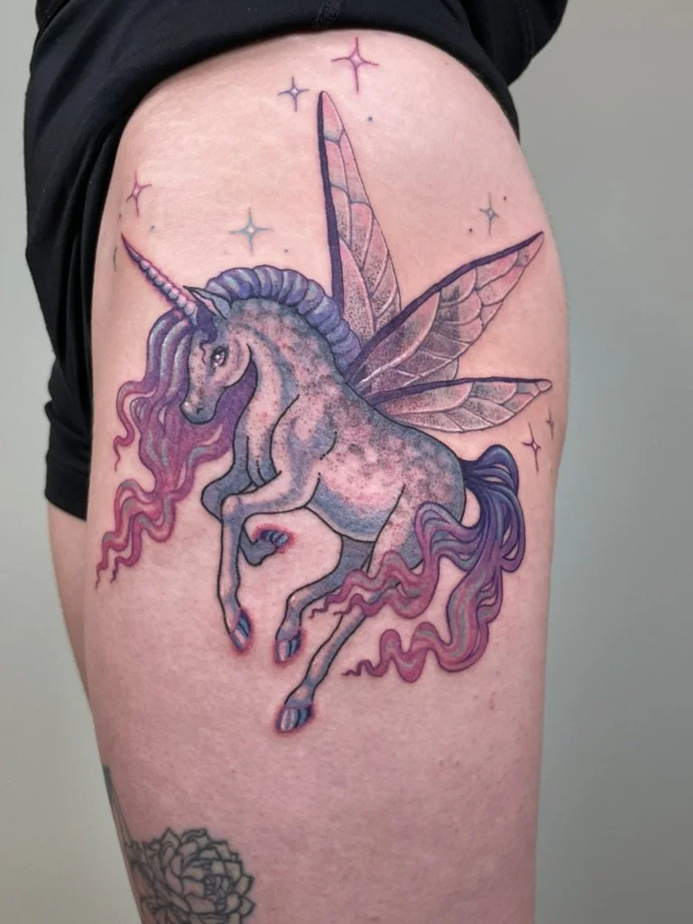Tattoo of a unicorn with fairy wings and a rainbow mane on a person's thigh.