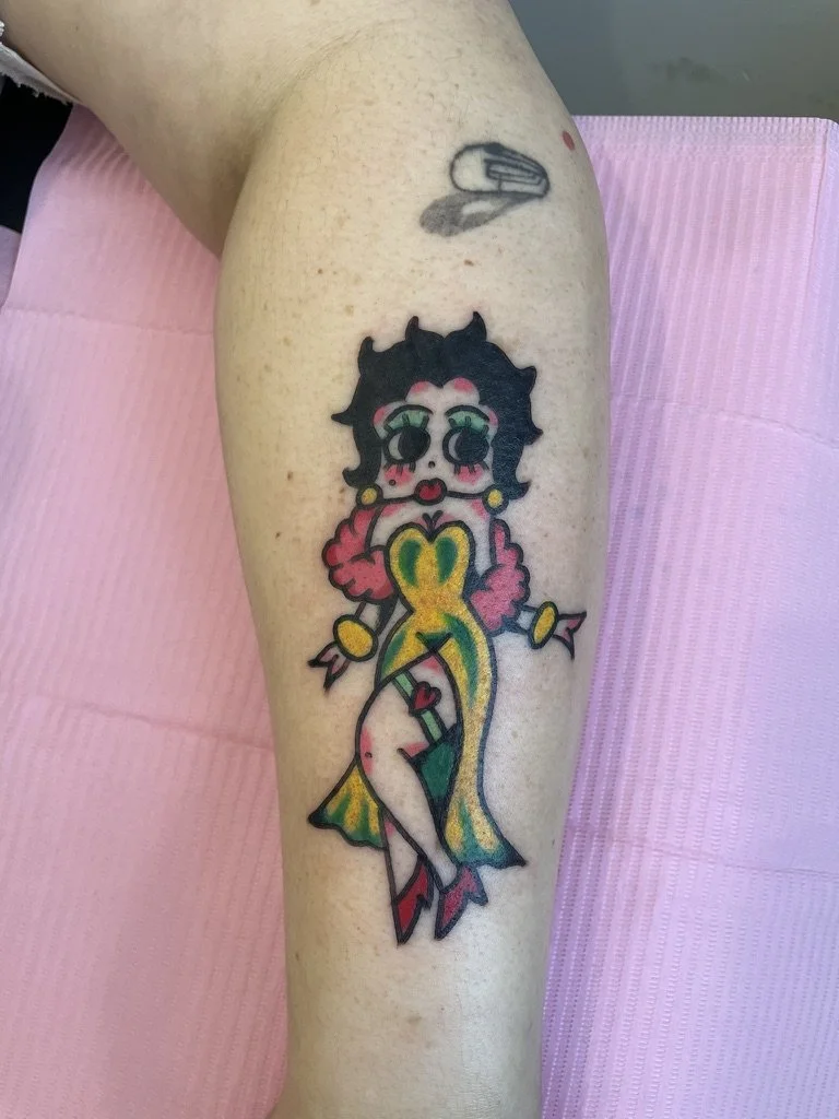Colorful tattoo of a cartoon woman with black hair, green eyes, pink cheeks, and red lipstick, wearing a yellow, pink, and green dress with a slit, pink earrings, and red high heels, on a person's leg.