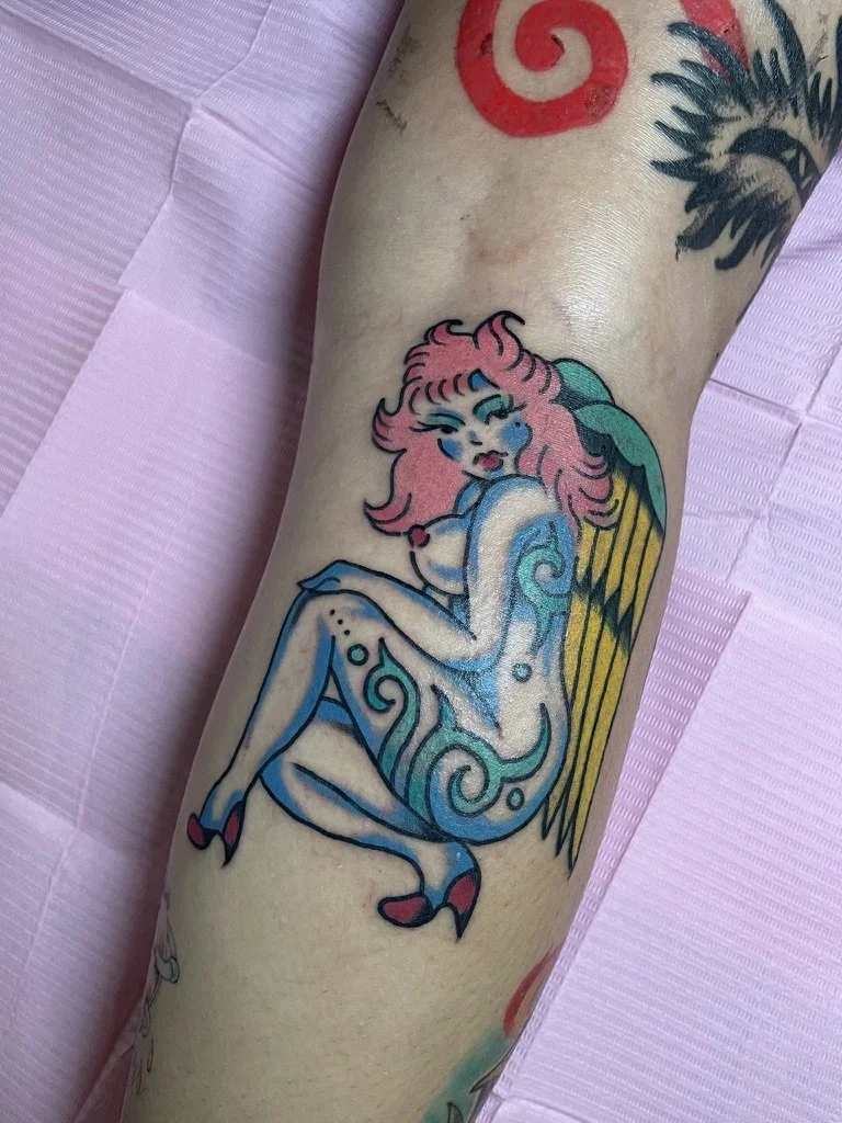 Tattoo of a mermaid with pink hair, blue skin, and colorful fins, sitting in a pose with her arms wrapped around her knees.