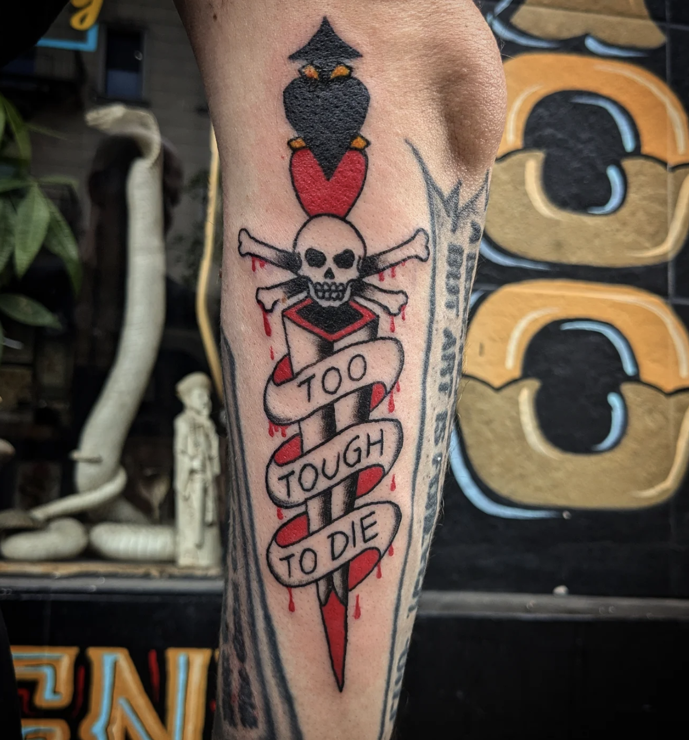 Tattoo of a sword with a skull and crossbones on top, wrapped with a banner that reads 'TOO TOUGH TO DIE,' with some red ink accenting the design.
