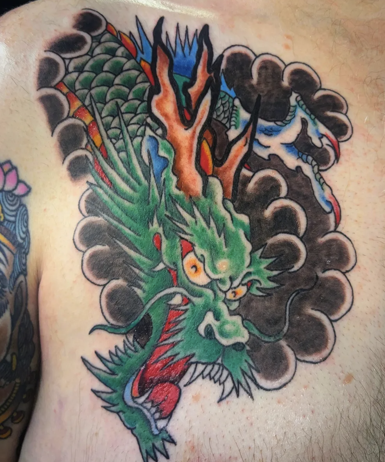 Colorful tattoo of a traditional Japanese style dragon with green scales, orange horns, and a fierce expression surrounded by dark clouds.