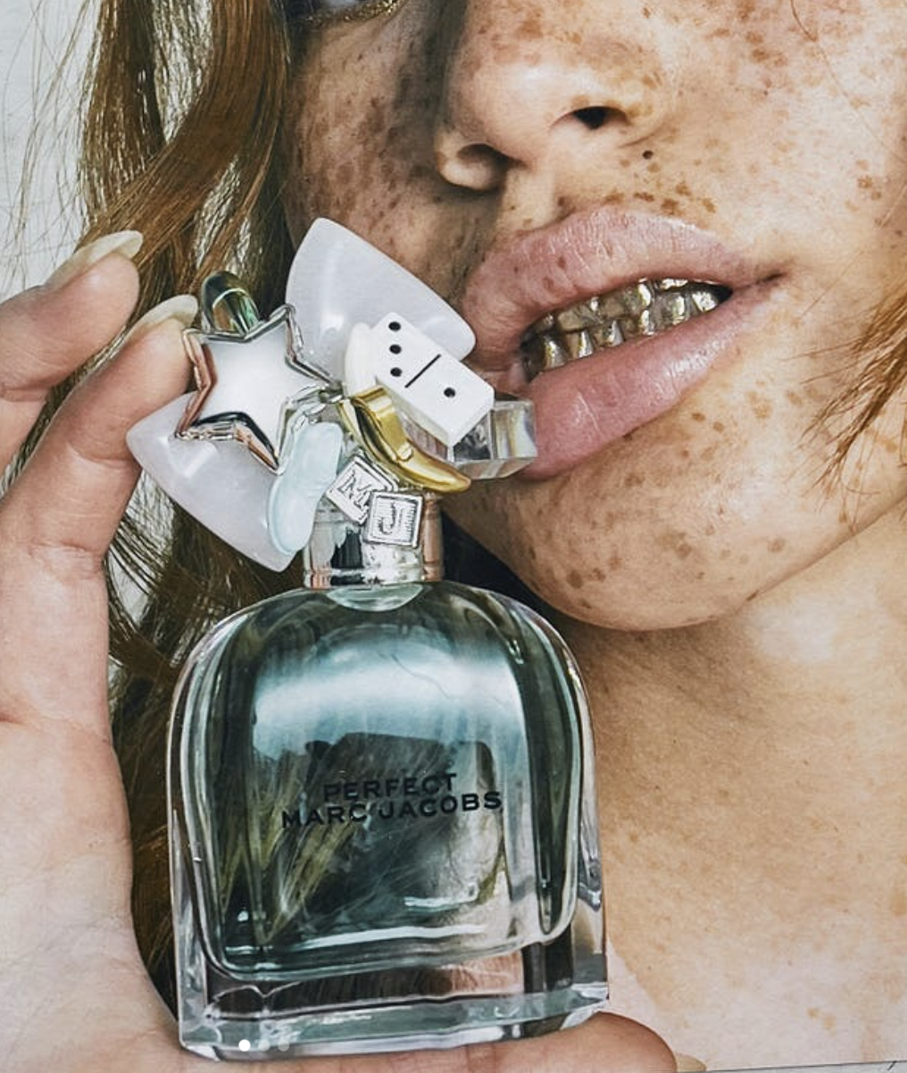 A woman with dark hair, wearing a black headband, is holding a bottle of perfume labeled 'Perfect Marc Jacobs' and playfully sticking out her tongue.