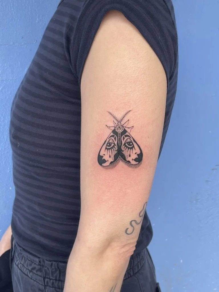 A tattoo of a moth with human eyes on its wings on a person's upper arm.