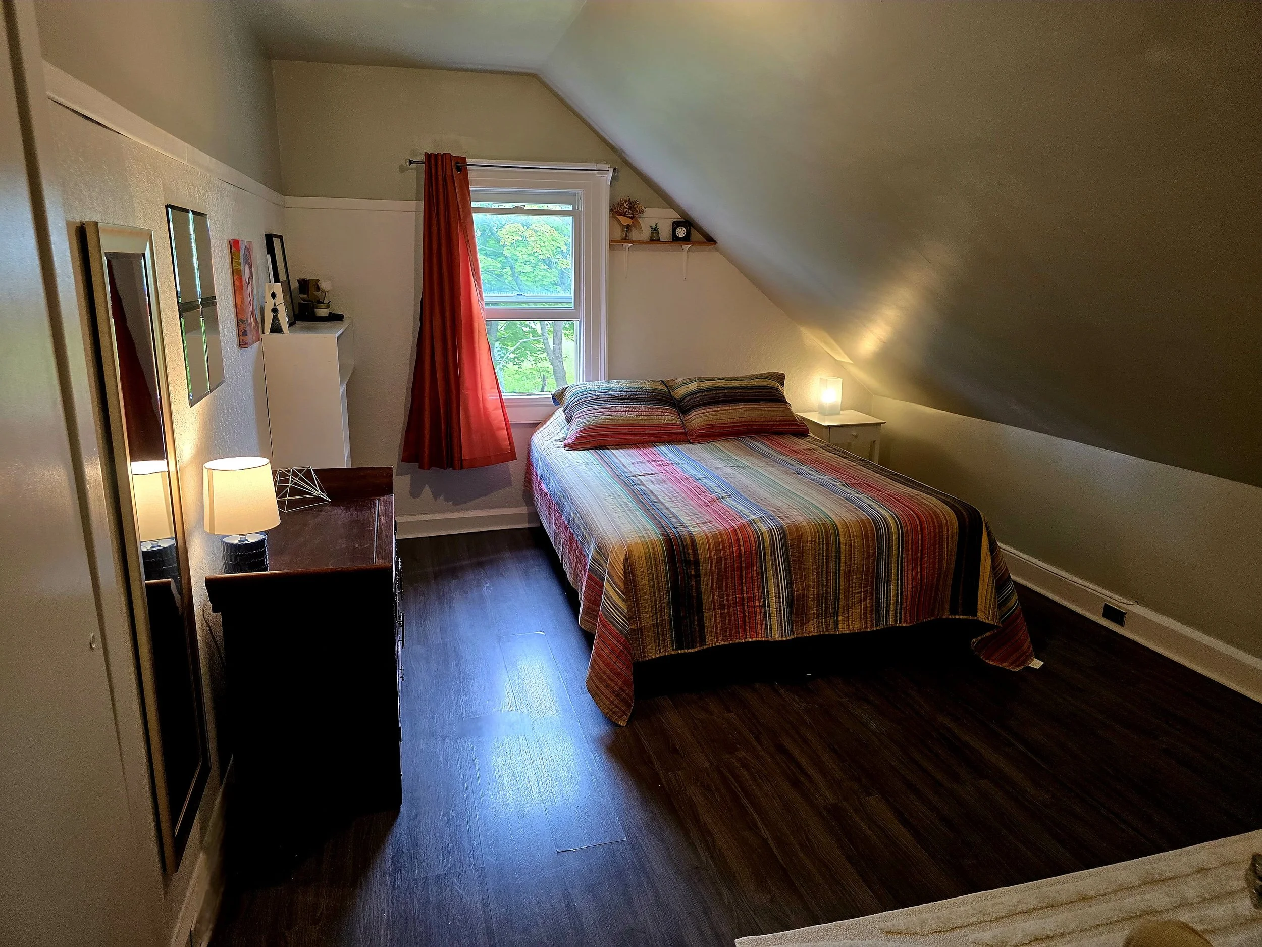 A bedroom with a bed covered in a colorful striped quilt, two pillows, a window with red curtains, a nightstand with a lamp, a dresser with lamps, and a small shelf with decor above the bed. The room has sloped ceilings and dark hardwood flooring.