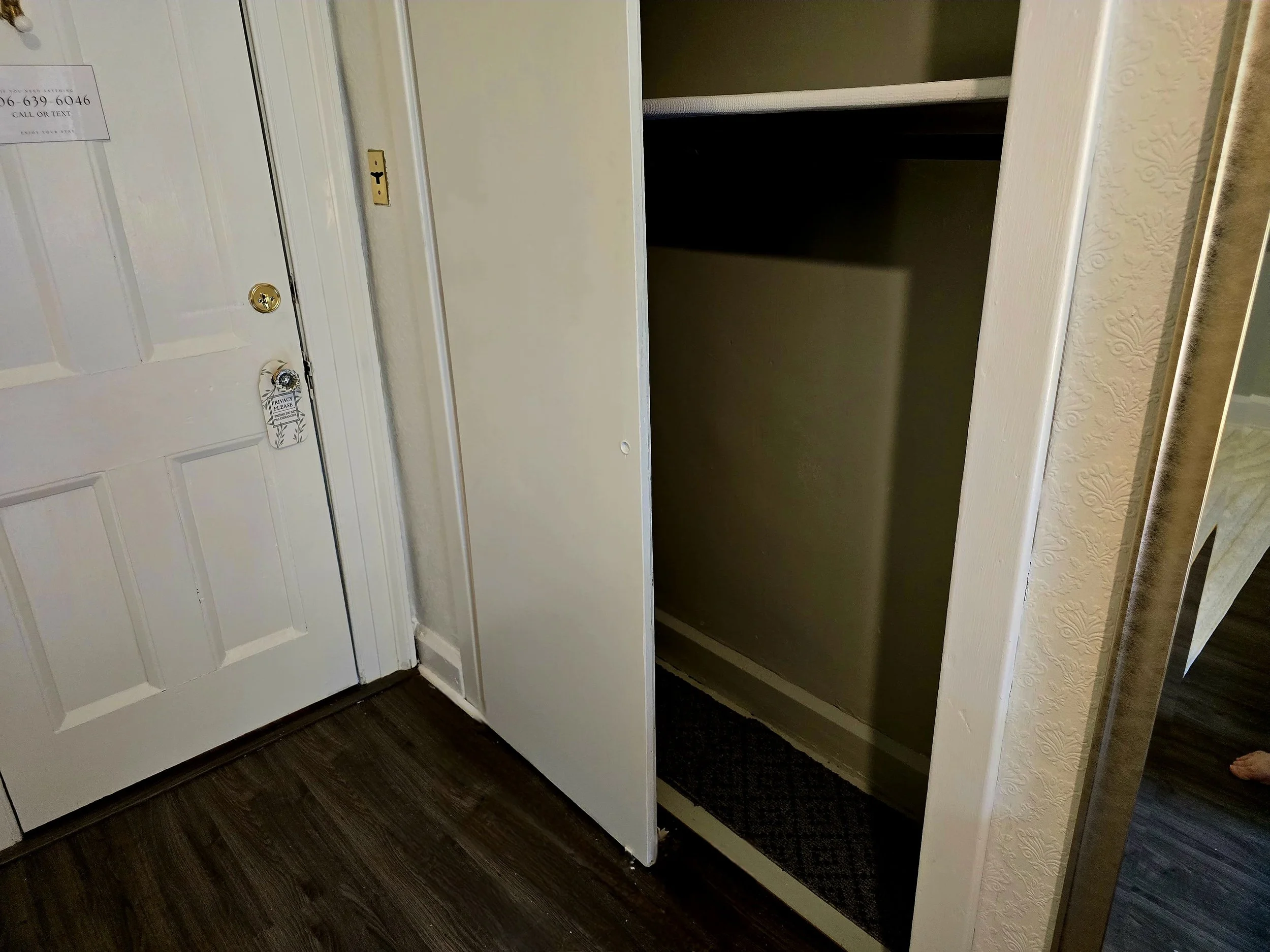 A closet with an open door showing an empty interior and a dark shelf inside. To the left, a white door with a jewelry tag hanging on it and a small sign posted near the top.