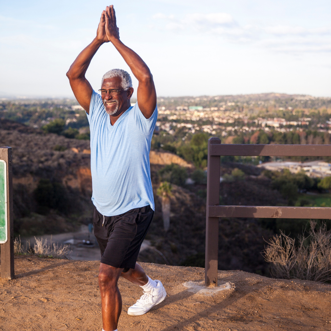 Older man with grey hair and glasses stretching outdoors on a trail with cityscape in the background, wearing a light blue t-shirt, black shorts, and white sneakers.