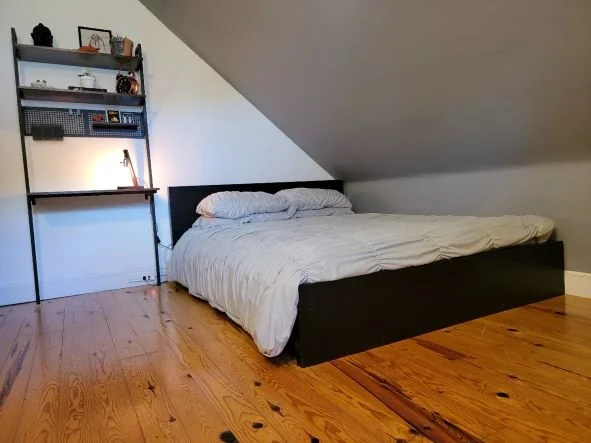 A bedroom with a bed against the sloped ceiling wall, with a black frame and light-colored bedding. To the left, there is a black desk with a lamp, shelves, and decorative items, all on a wooden floor.