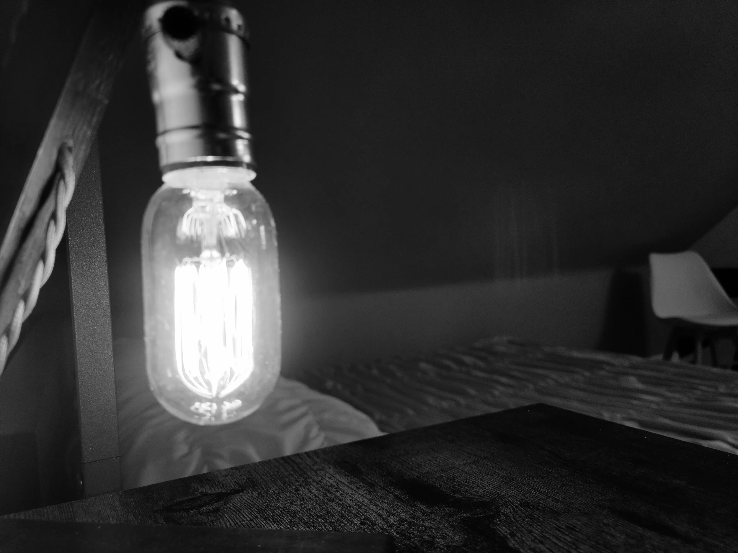 Close-up of a glowing Edison-style light bulb hanging from a ceiling, casting shadows on a nearby bed and furniture in a dark room.