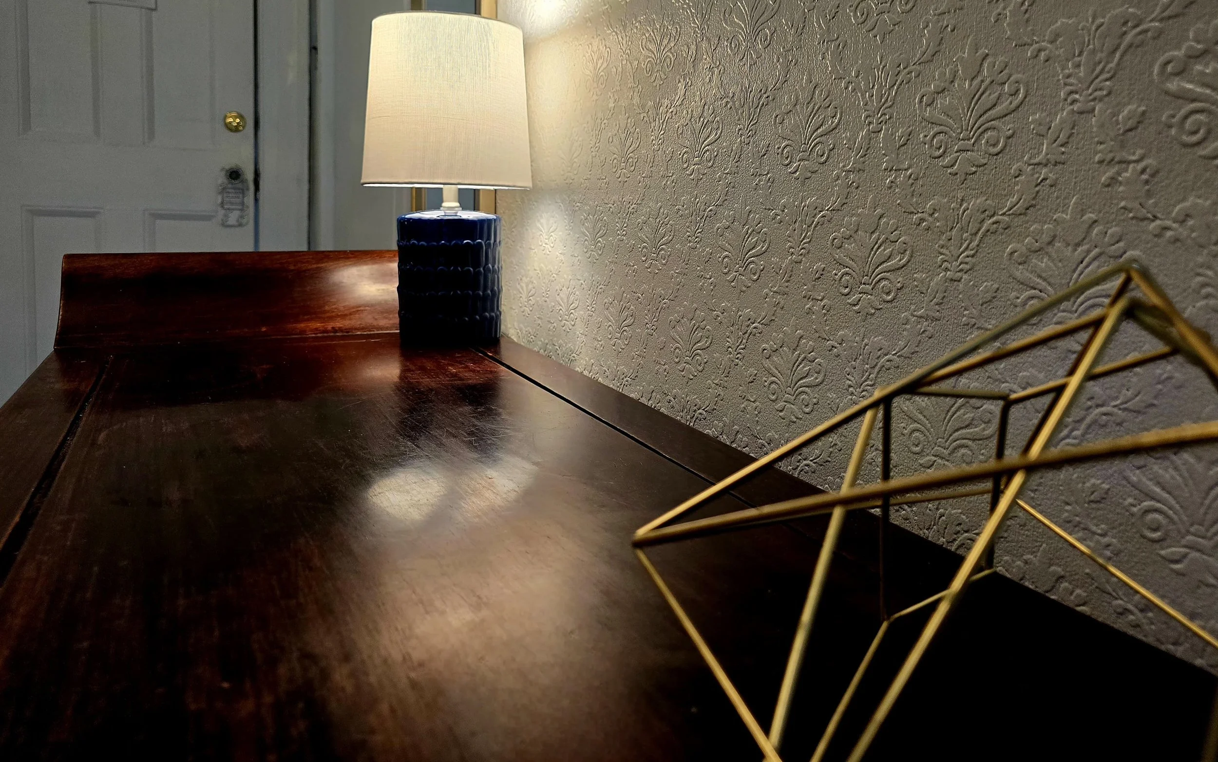 Close-up of a wooden desk with a table lamp, a decorative gold geometric object, and textured wallpaper in the background.