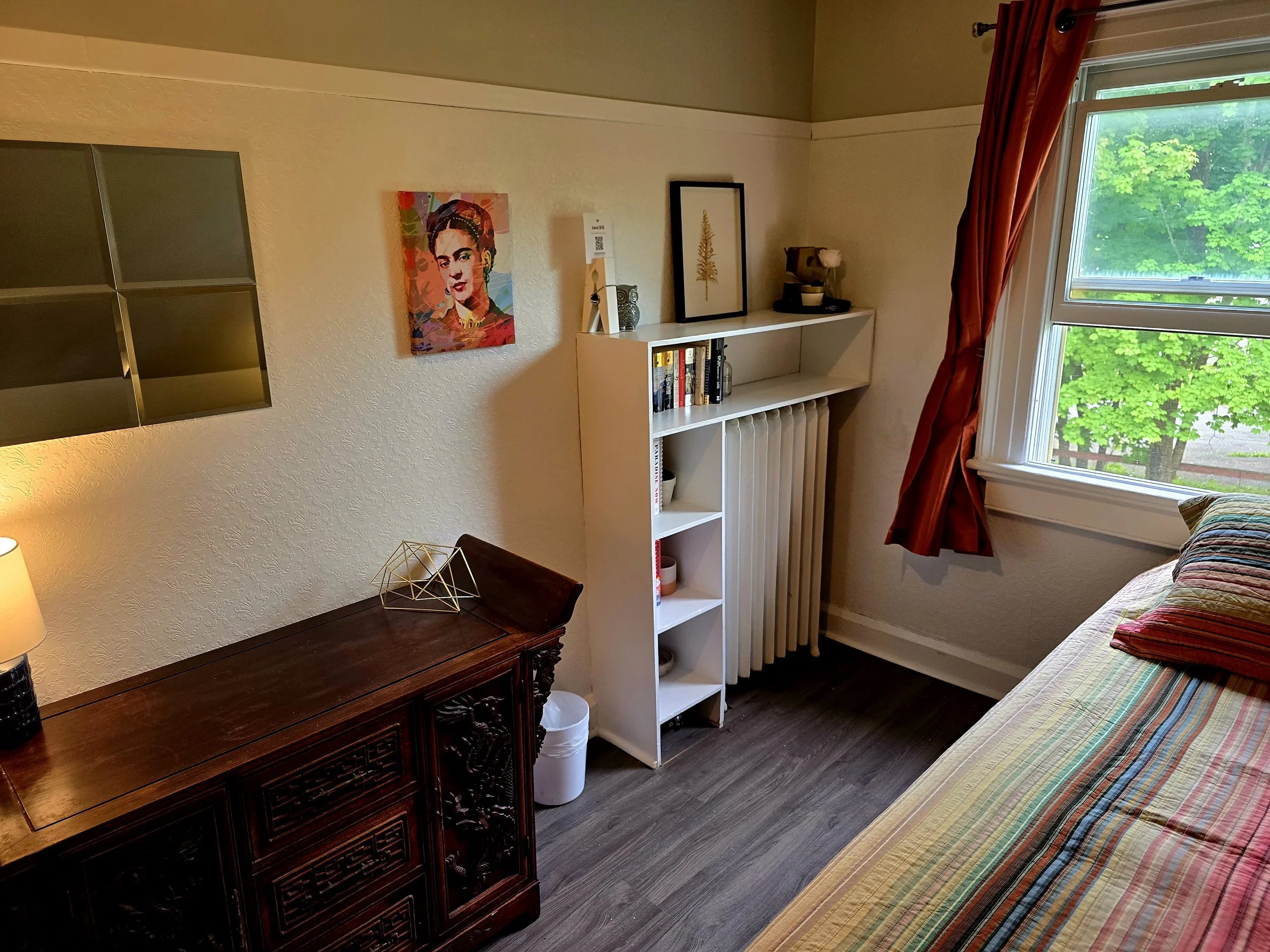 A cozy bedroom corner with a window showing green trees outside, a bed with striped bedding, red curtains, a white bookshelf with books and decorative items, a dark wooden dresser, and colorful artwork on the wall.