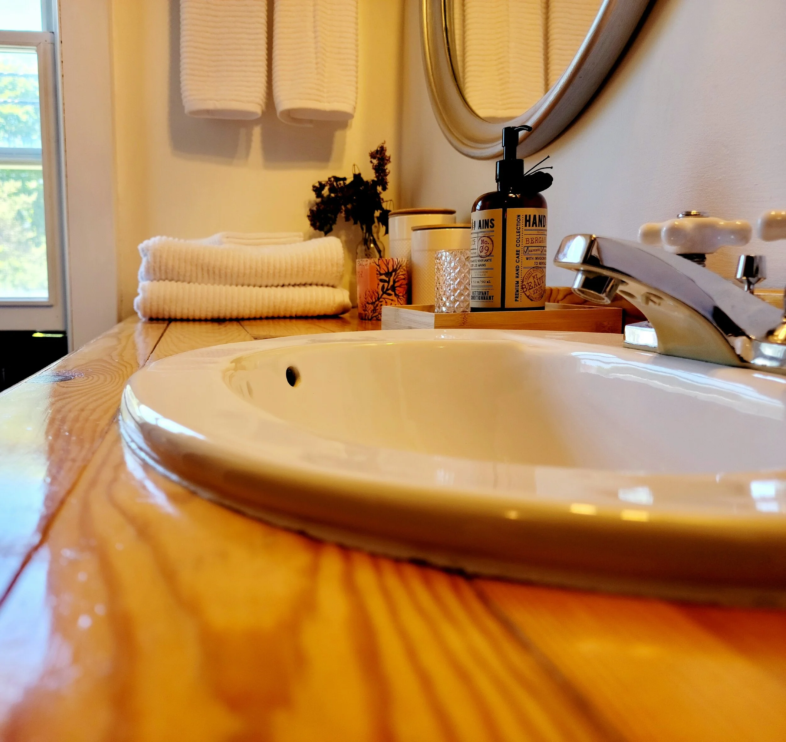 Close-up of a bathroom sink with a wooden countertop, in a cozy bathroom decorated with towels, a soap dispenser, candles, and a mirror.