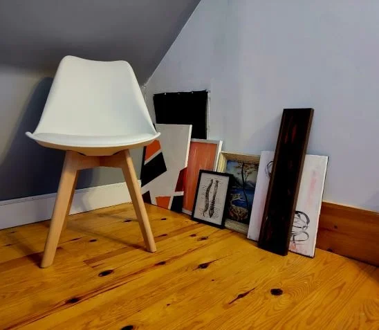 A white modern chair with wooden legs placed on a wooden floor, leaning against a sloped wall with several framed pictures stacked on the floor beside it, in an attic or corner space.