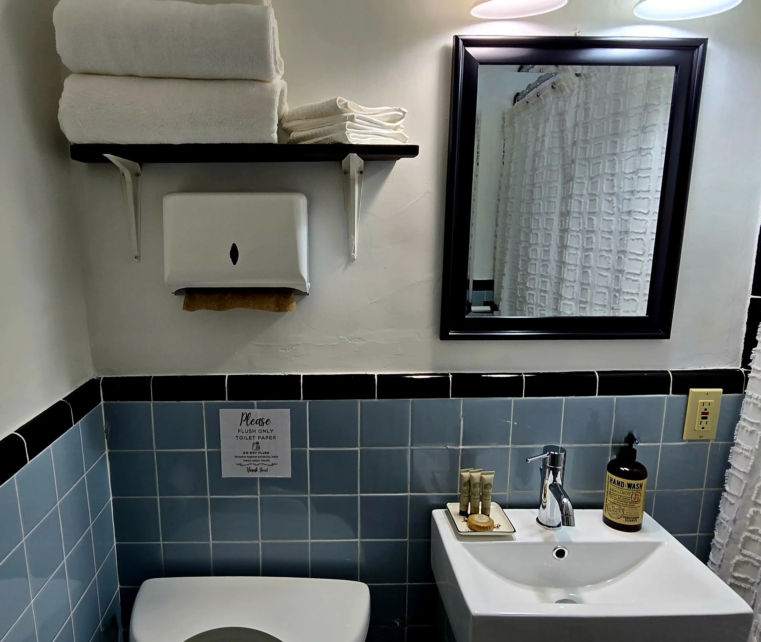 A bathroom scene showing a mirror, a soap dispenser, a sink with toiletries, a towel, and a toilet, with a shower curtain reflected in the mirror.