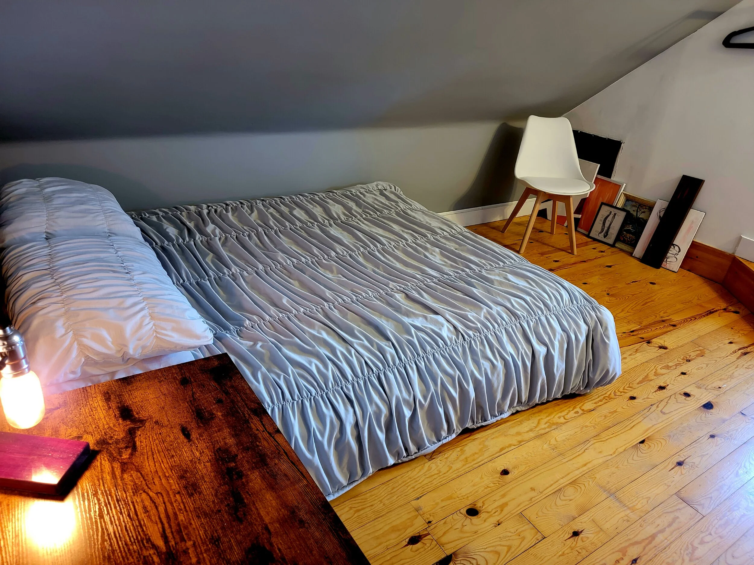 A bedroom with a bed covered in a silver-colored quilt, a pillow, a wooden nightstand with a light bulb, and a white chair. Several framed pictures and artwork are stacked near the wall on the floor. The room has hardwood floors and a sloped ceiling.