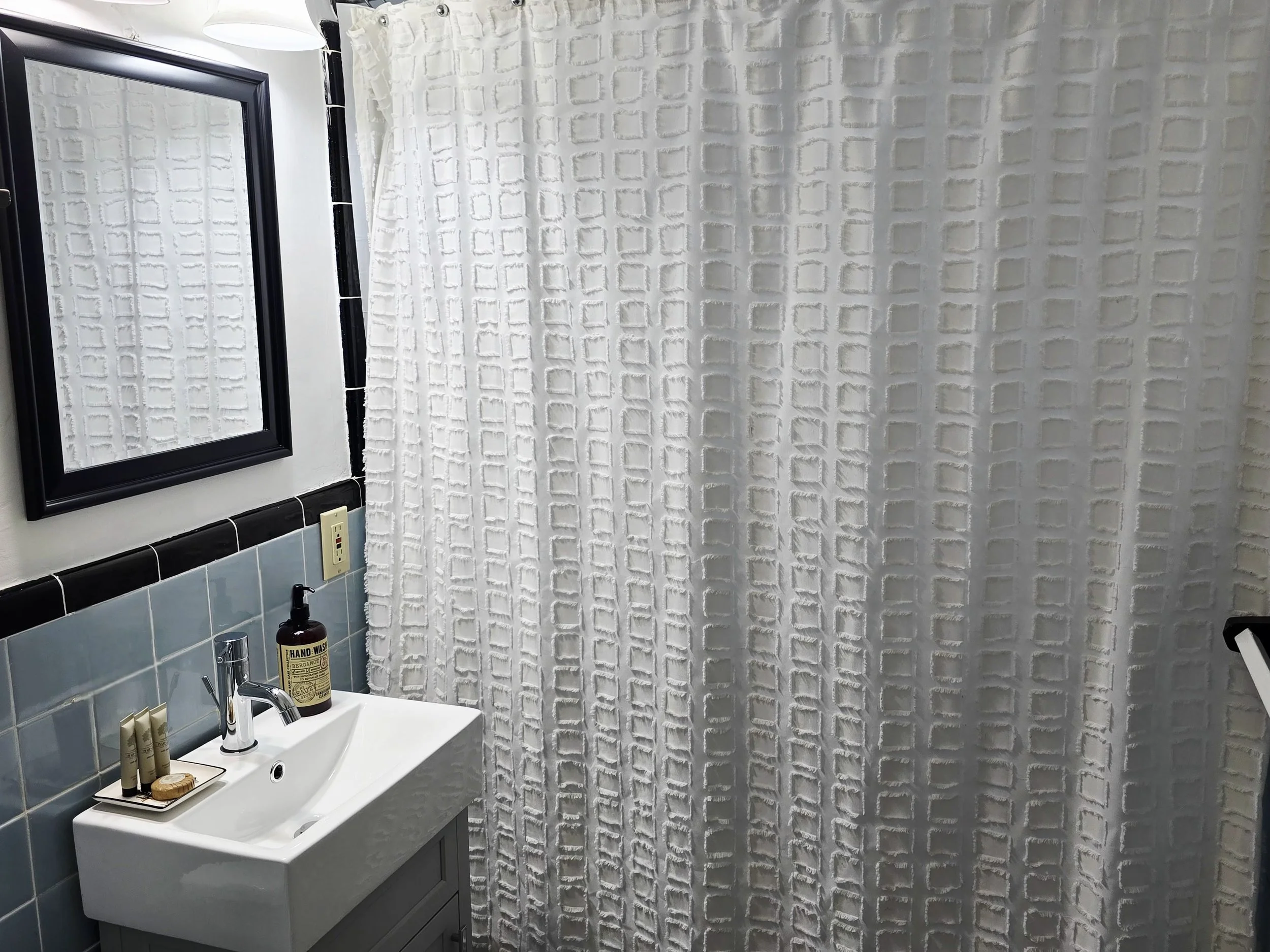 Bathroom with white textured shower curtain, mirror with black frame, small sink with blue tiled backsplash, and hand soap on the sink.