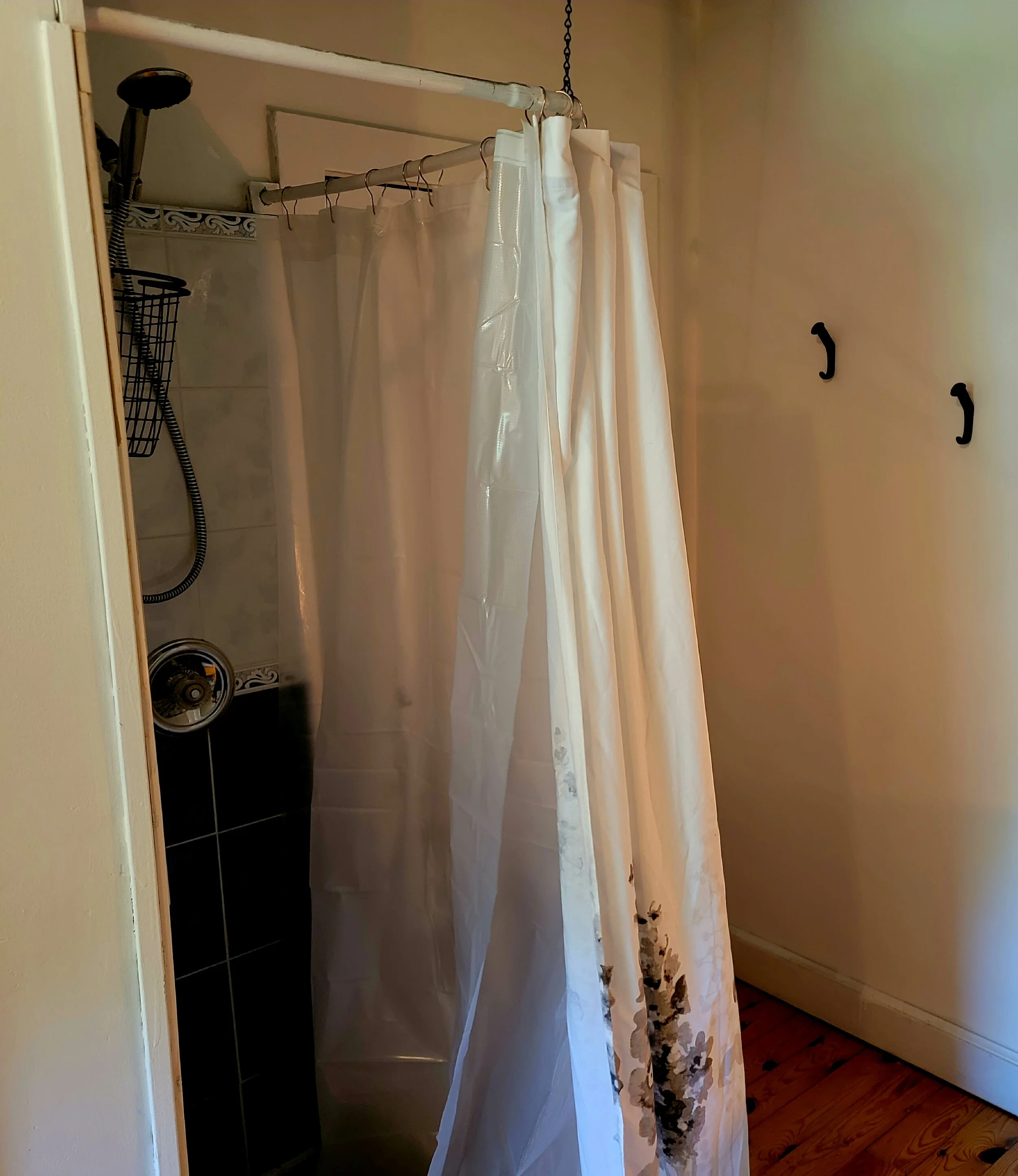 Bathroom shower with white curtain and dark tiles, showerhead, and wall hooks.