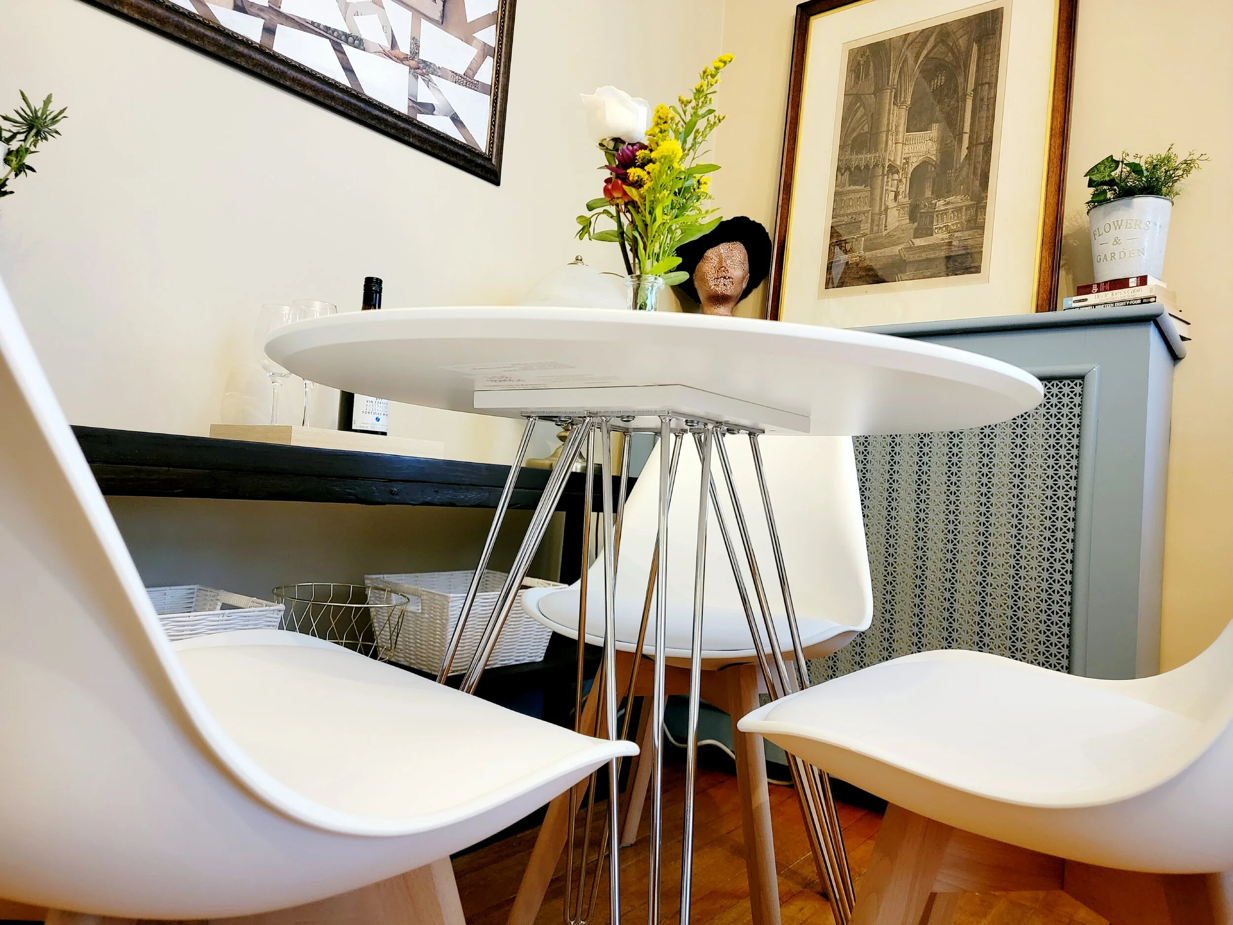 A dining room with a white round table, two white chairs, a gray cabinet, framed artwork, and decorative plants.