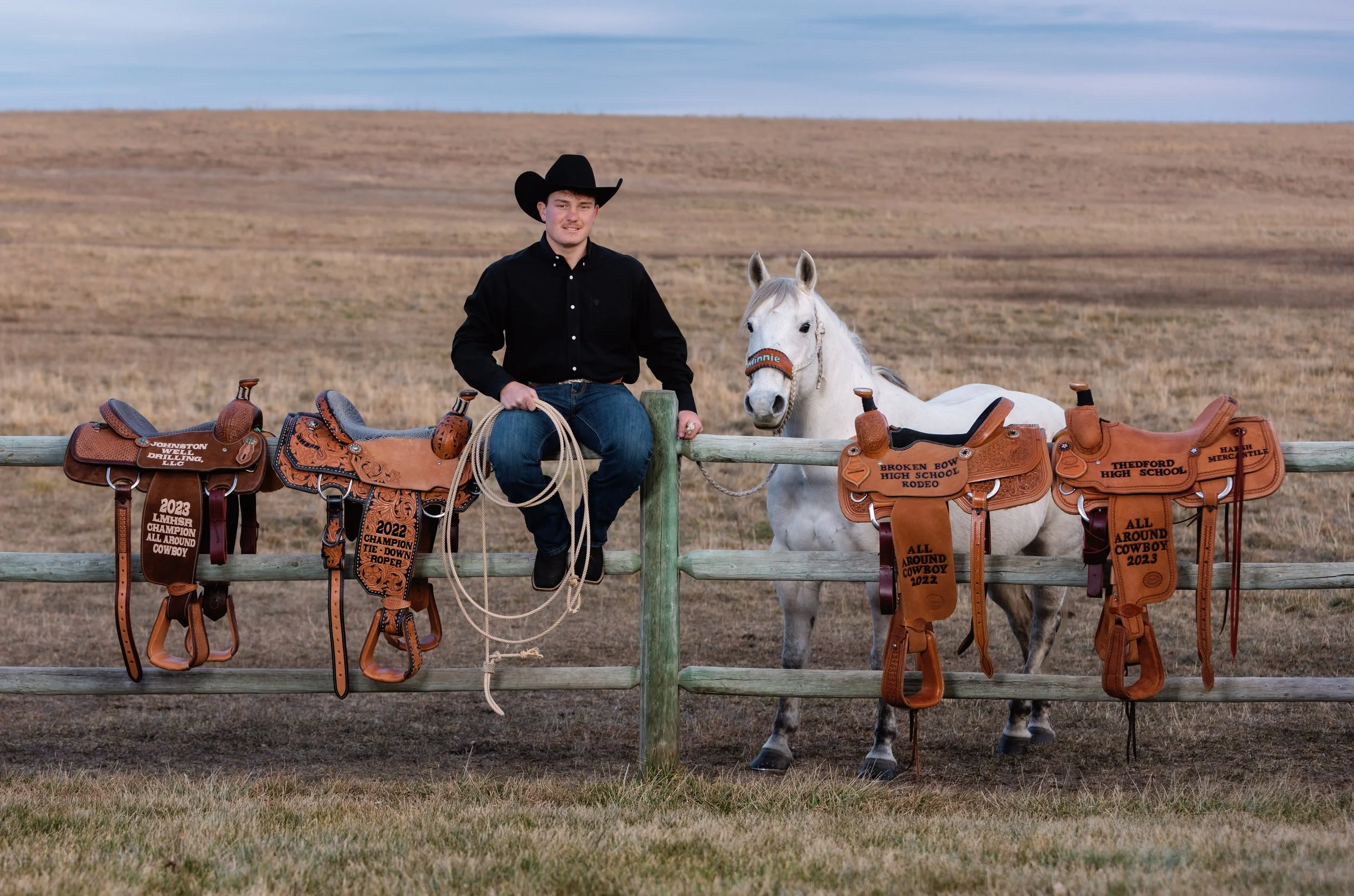 Central nebraska high school senior Sin with his grey horse sitting on fence with four award saddles