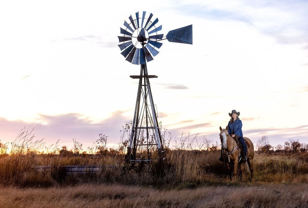 palomino horse and lady rider with a gray hat by a stock tank and windmill at sunset in central nebraska