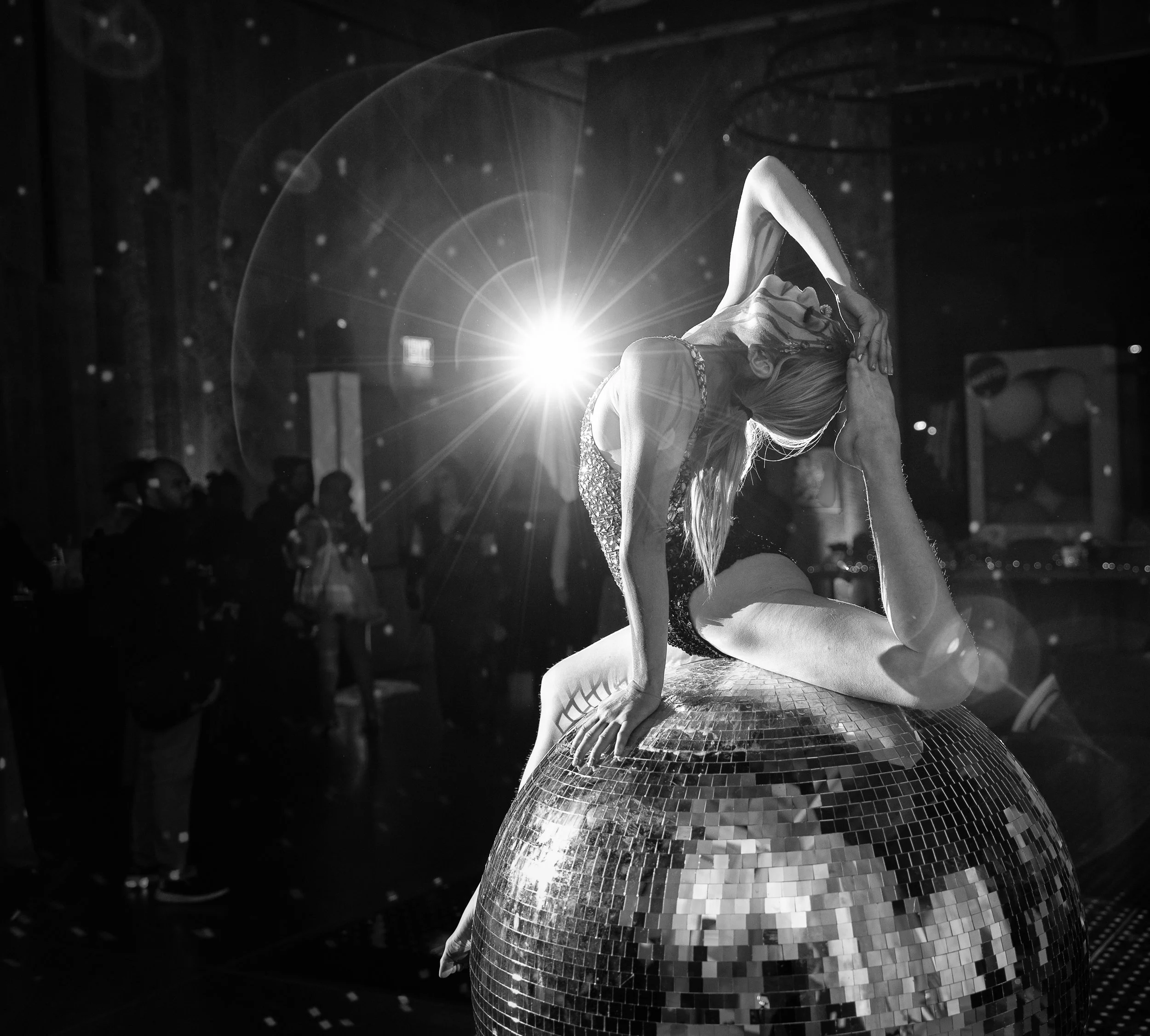 A woman in a dance pose, sitting on a large disco ball, illuminated by bright stage lights in a dark venue with an audience in the background.