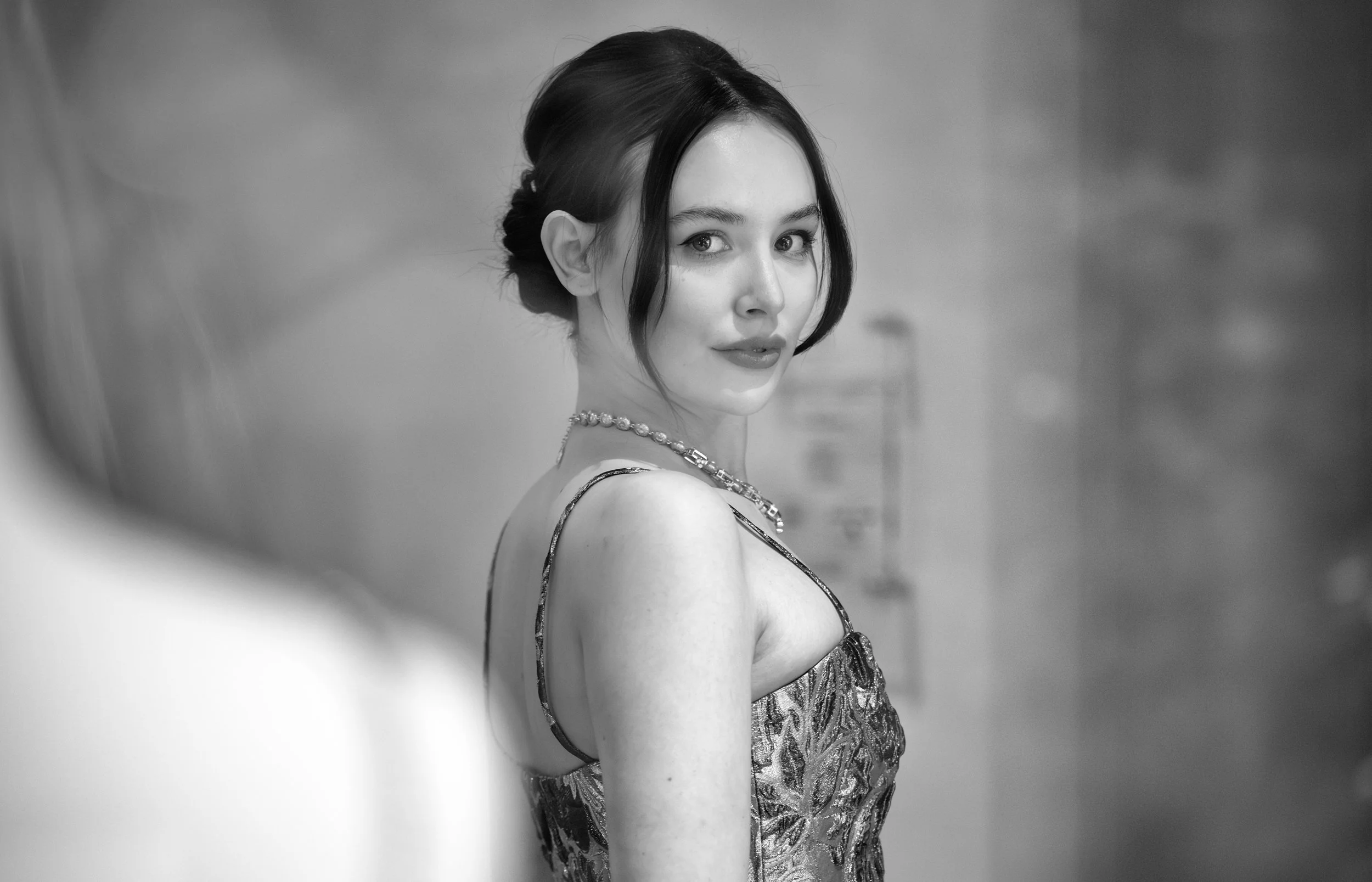 A woman with dark hair styled in an elegant updo, wearing a patterned dress and a pearl necklace, smiling softly while looking at the camera. The background is blurred.
