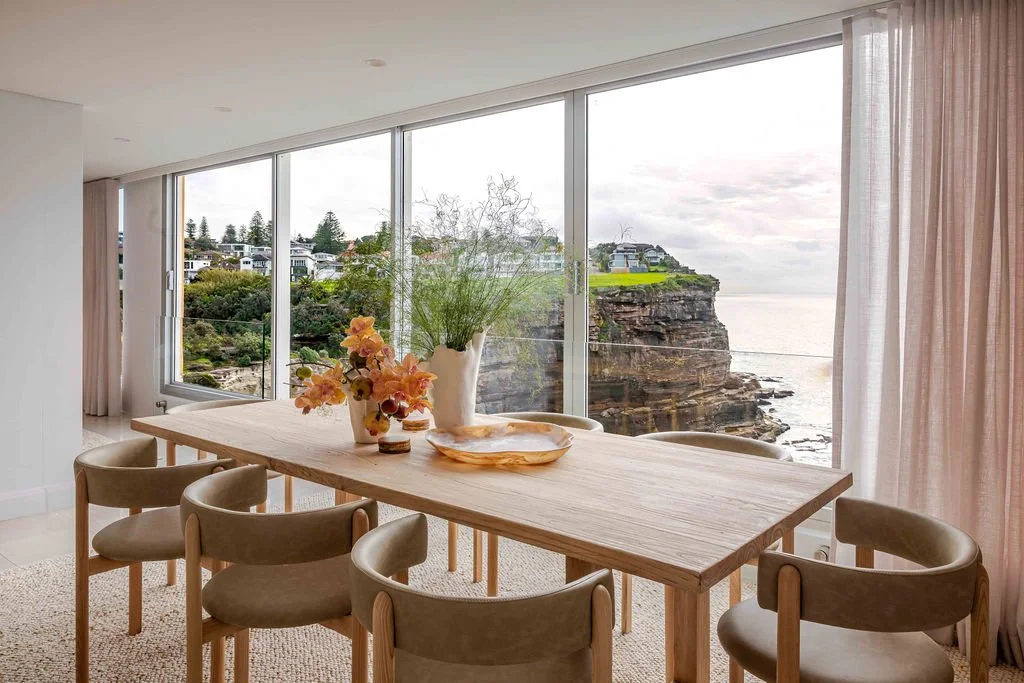 A dining room with a large wooden table and six beige chairs, decorated with vases of flowers, situated next to floor-to-ceiling windows overlooking a rocky coastline, ocean, and houses on a cliff.