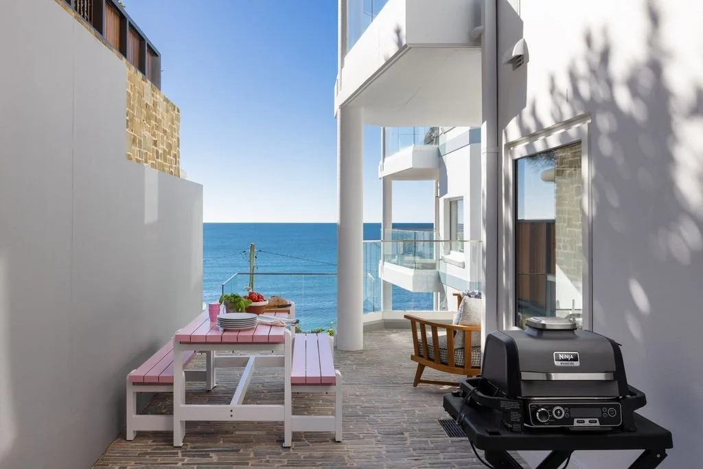 Balcony with a pink picnic table set for outdoor dining, a charcoal grill, and a sea view with balconies of neighboring buildings.