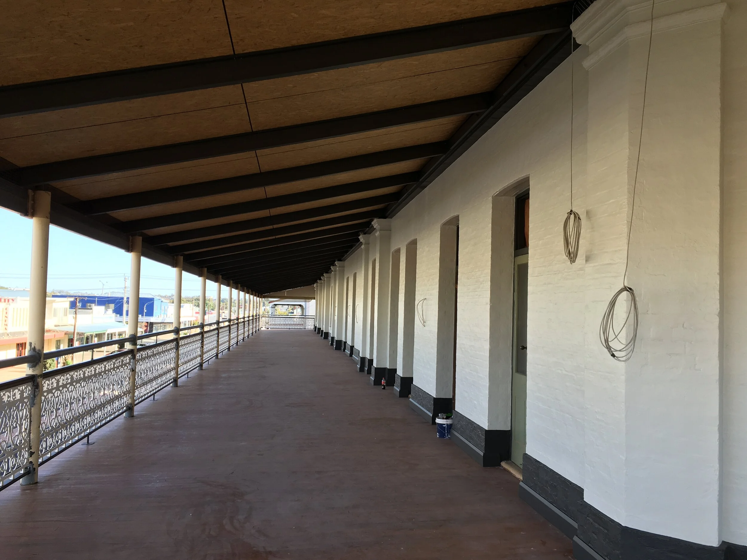 An outdoor balcony with white brick walls, black baseboards, and a wooden ceiling, temporarily under construction with hanging wires and paint buckets along the wall.