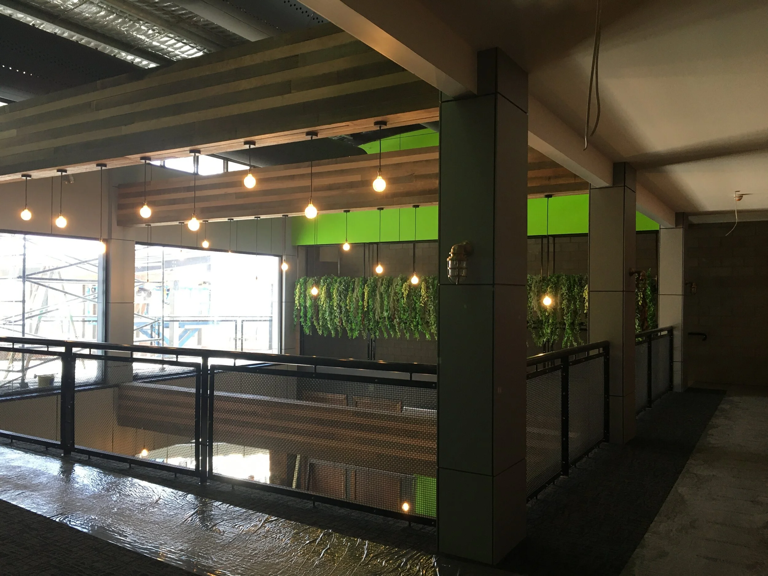 Interior view of an indoor space with hanging light bulbs, green accents, and decorative plants, overlooking a lower level through a safety railing.