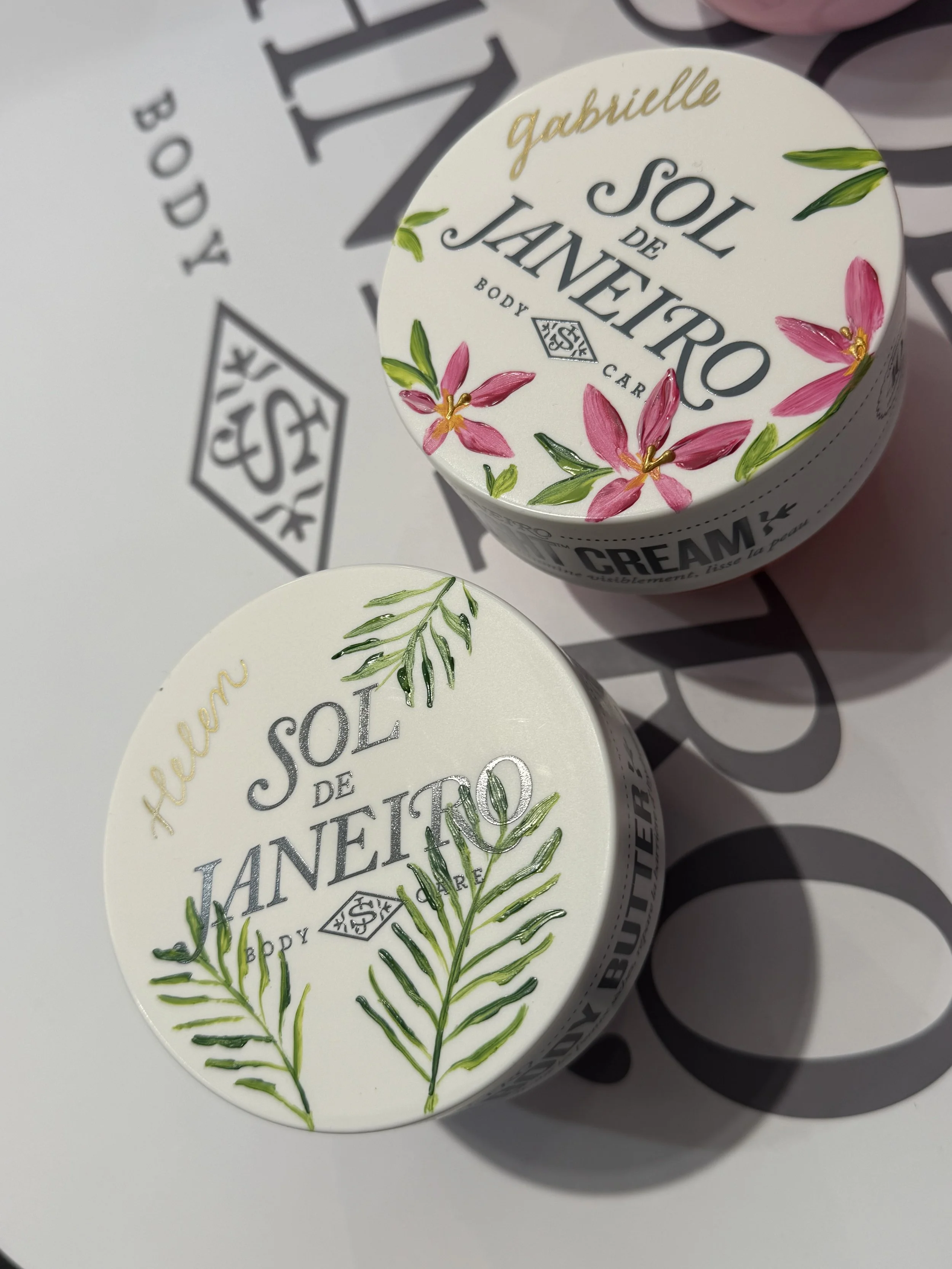 2 body creams painted. Left side, green palm leaves with name in gold calligraphy. Right side, red flowers with green leaves and name in gold calligraphy