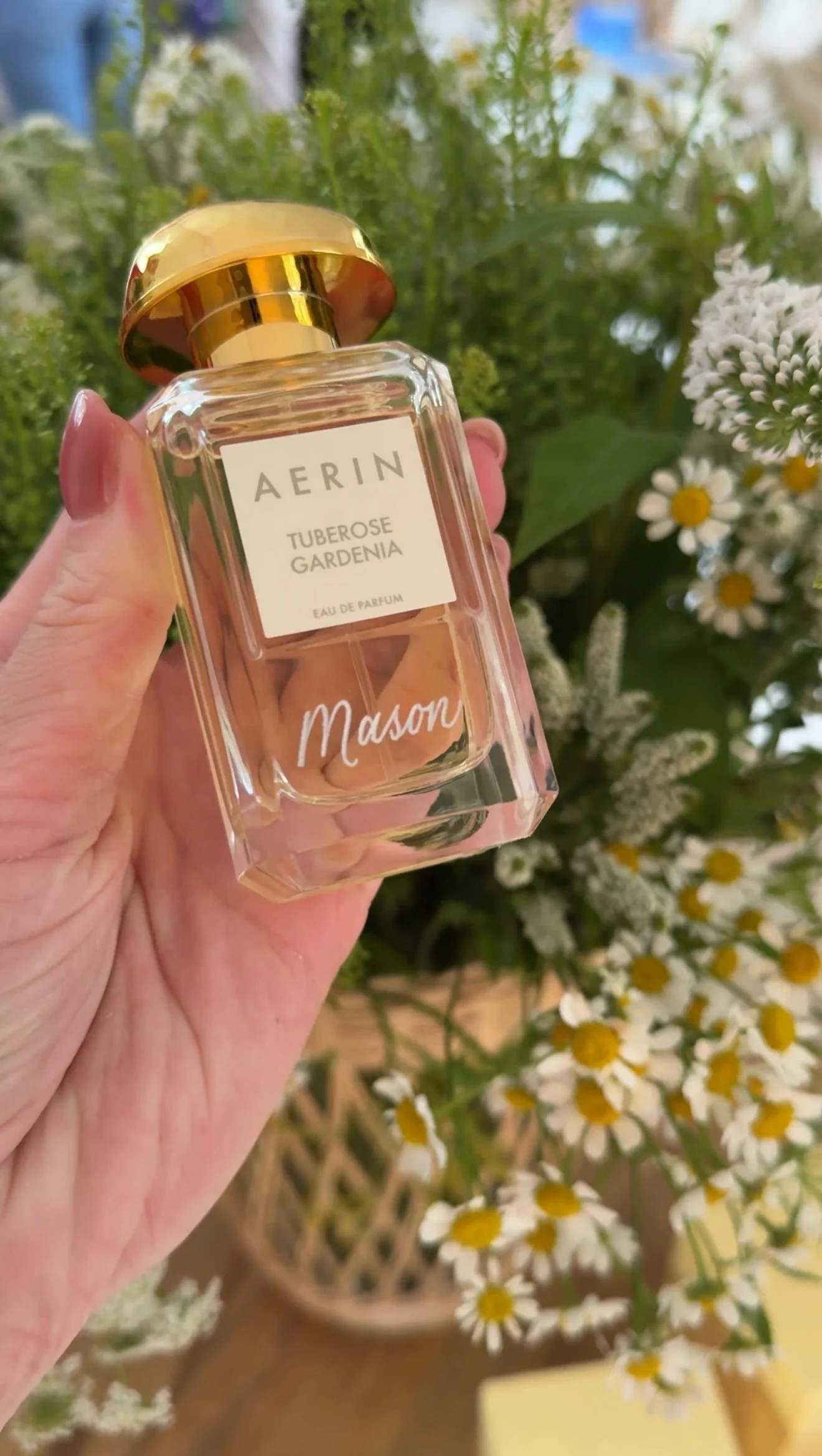 AERIN perfume bottle engraved in silver with the name Mason being held in front of a floral bouquet