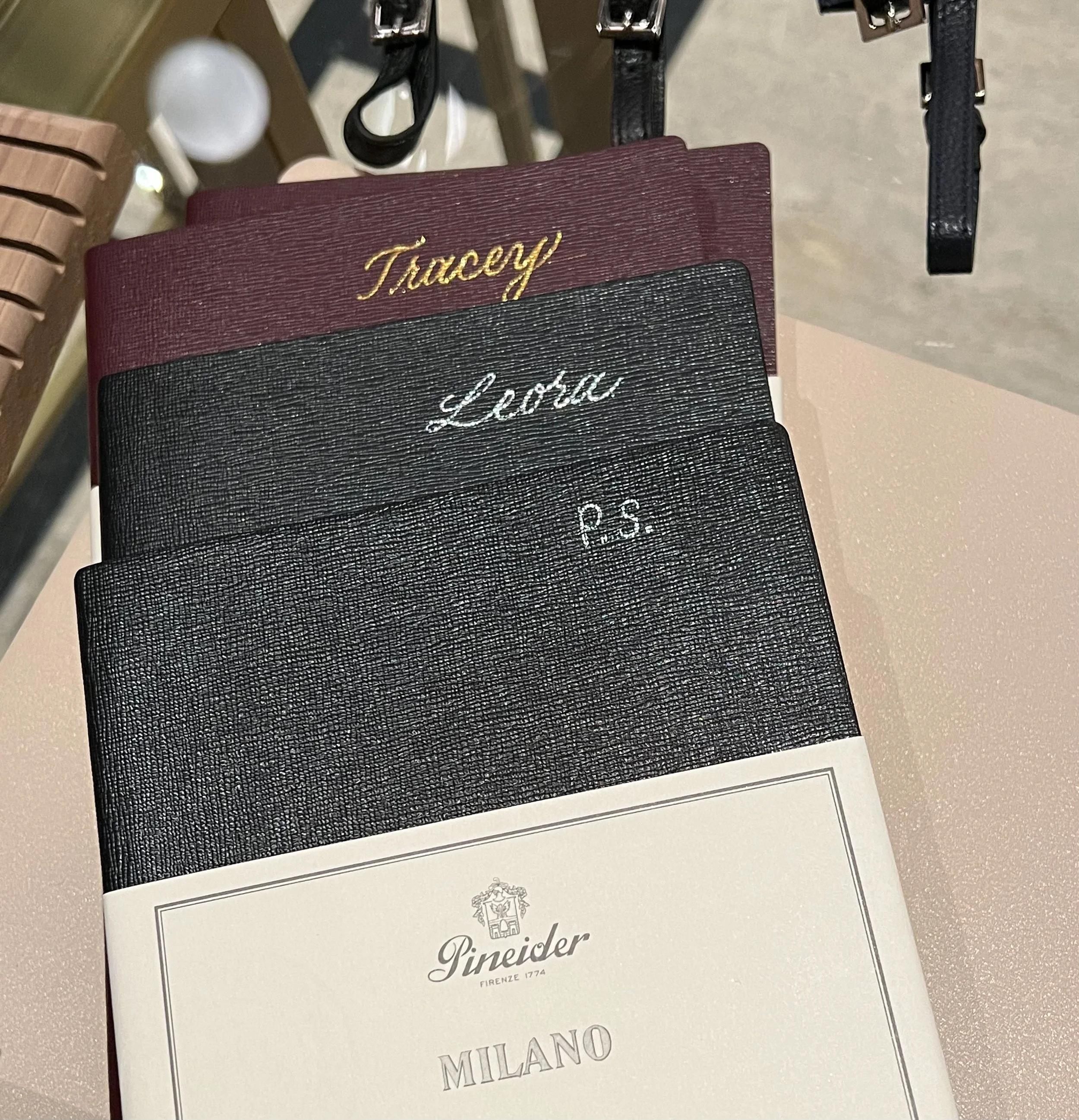 3 Pineider notebooks customized. Top Burgundy notebook personalized with Tracey in gold, then a black notebook with Leora in silver, and a black notebook with silver initials all caps P.S.