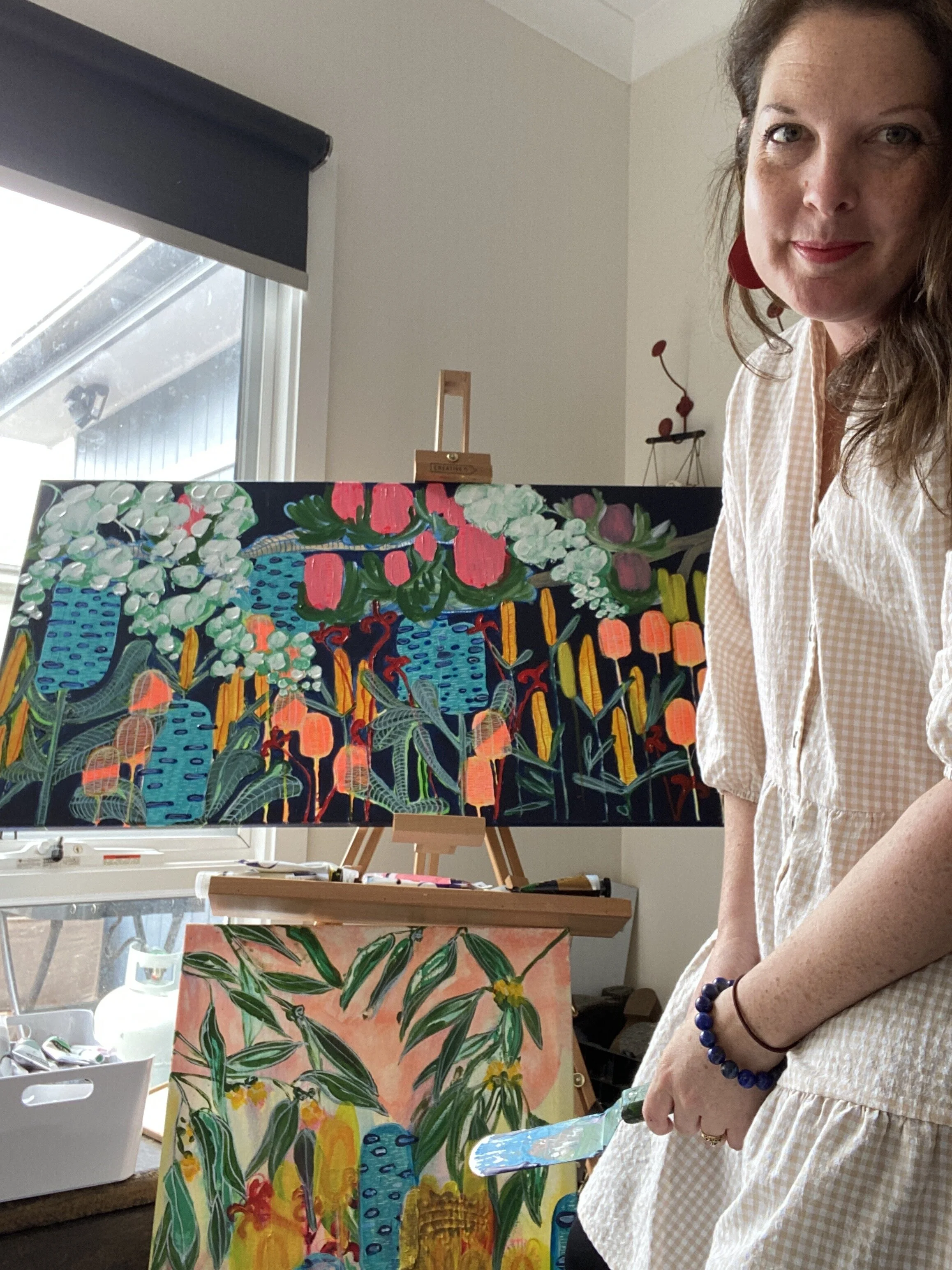 Woman standing next to two colorful paintings on easels in a bright room.