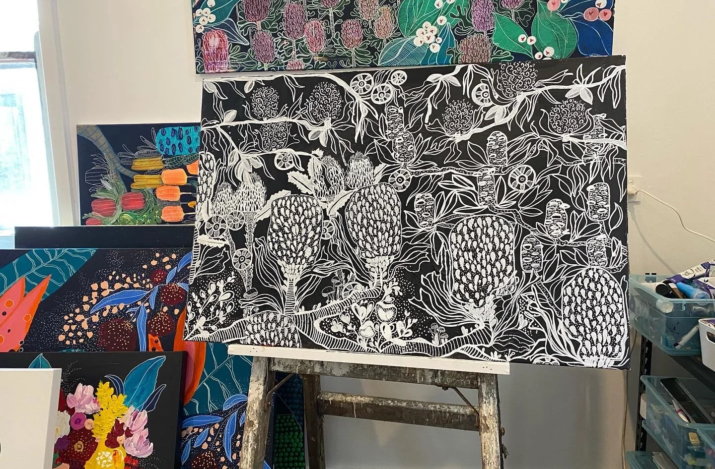 Black and white painting of abstract plants and flowers on an easel, with other colorful floral paintings partially visible behind it.