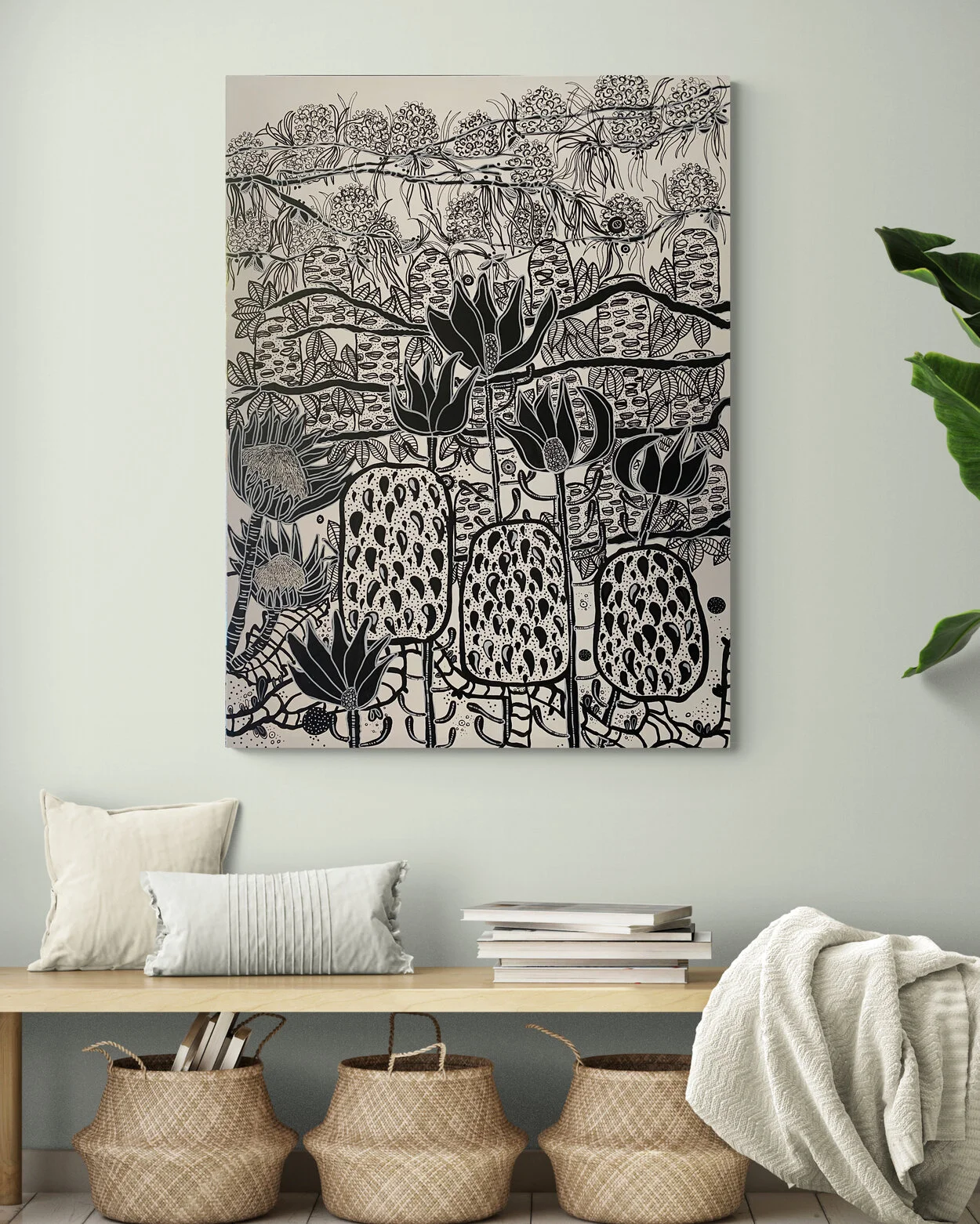 Decorative black and white botanical artwork hanging on a white wall in a room with a light-colored table, pillows, a stack of books, and woven baskets.