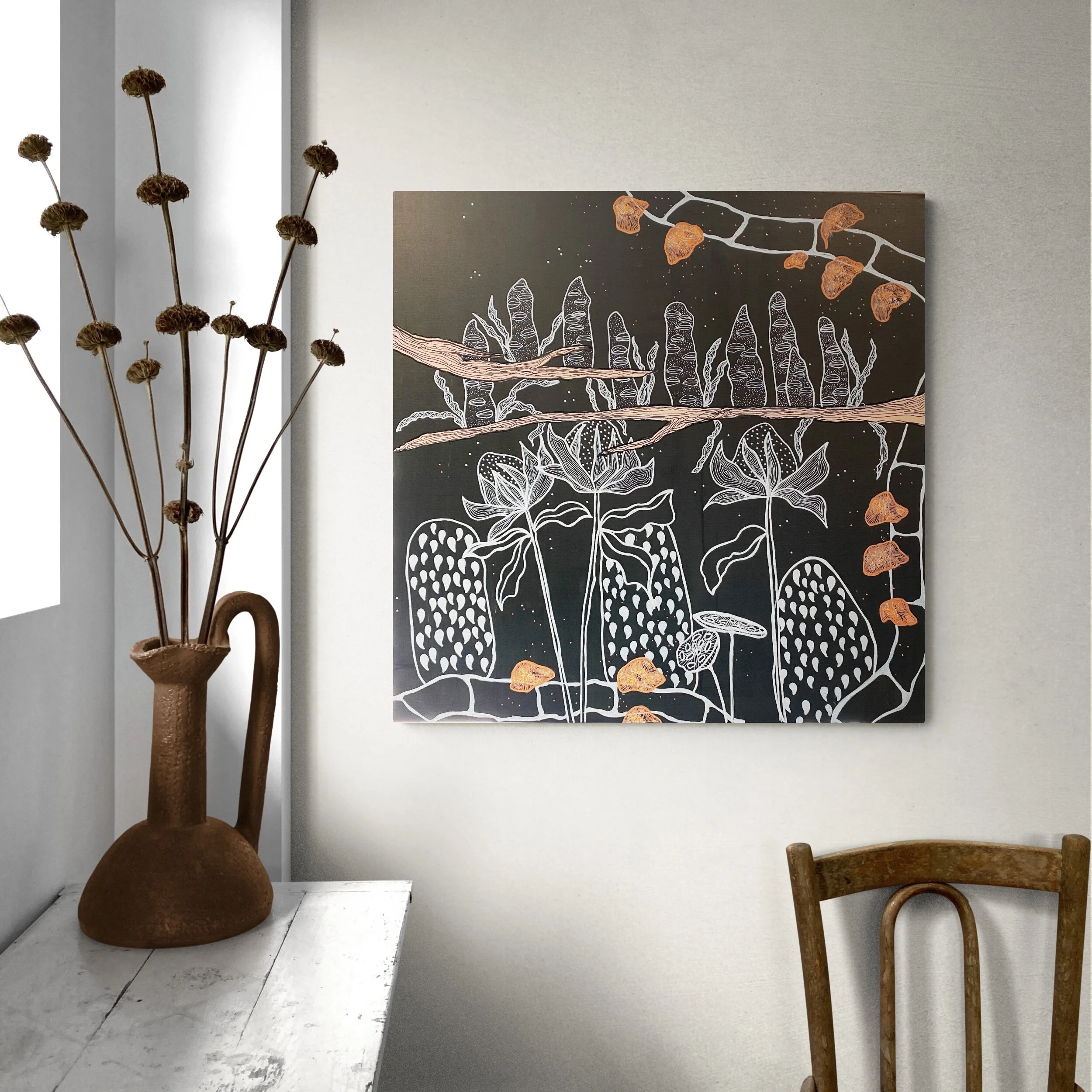 Room with abstract botanical artwork on wall, featuring white and orange flower patterns. A rustic brown vase with dried flowers sits on a white wooden table, next to a wooden chair.