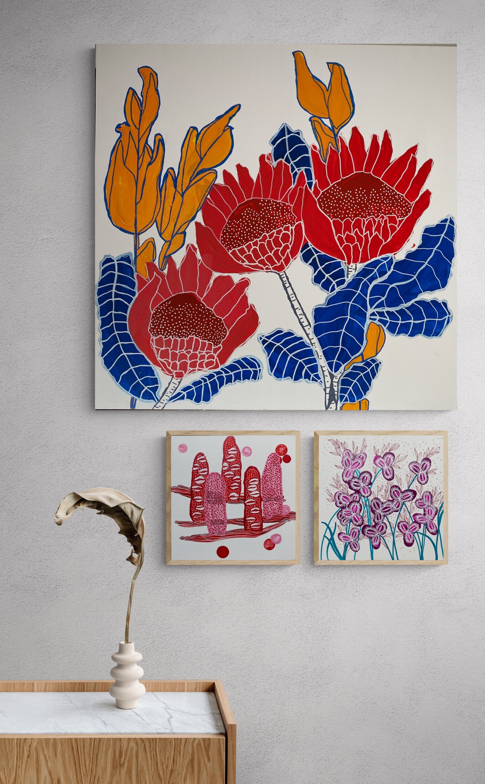 Three abstract floral paintings on a wall above a wooden console with a vase holding a single dried leaf.