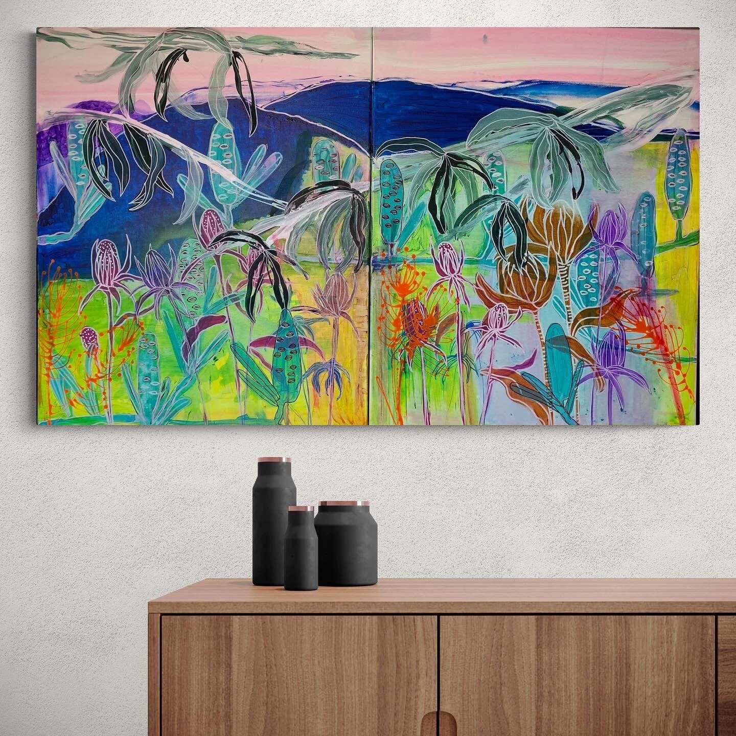 Abstract painting with colorful flowers and plants against a mountain background, displayed above a wooden cabinet with three black vases.