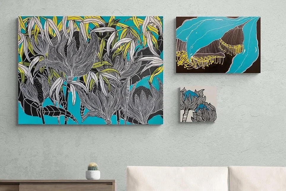 Three abstract paintings with botanical and abstract blue patterns on a light-colored wall, above a wooden surface with a small cactus.