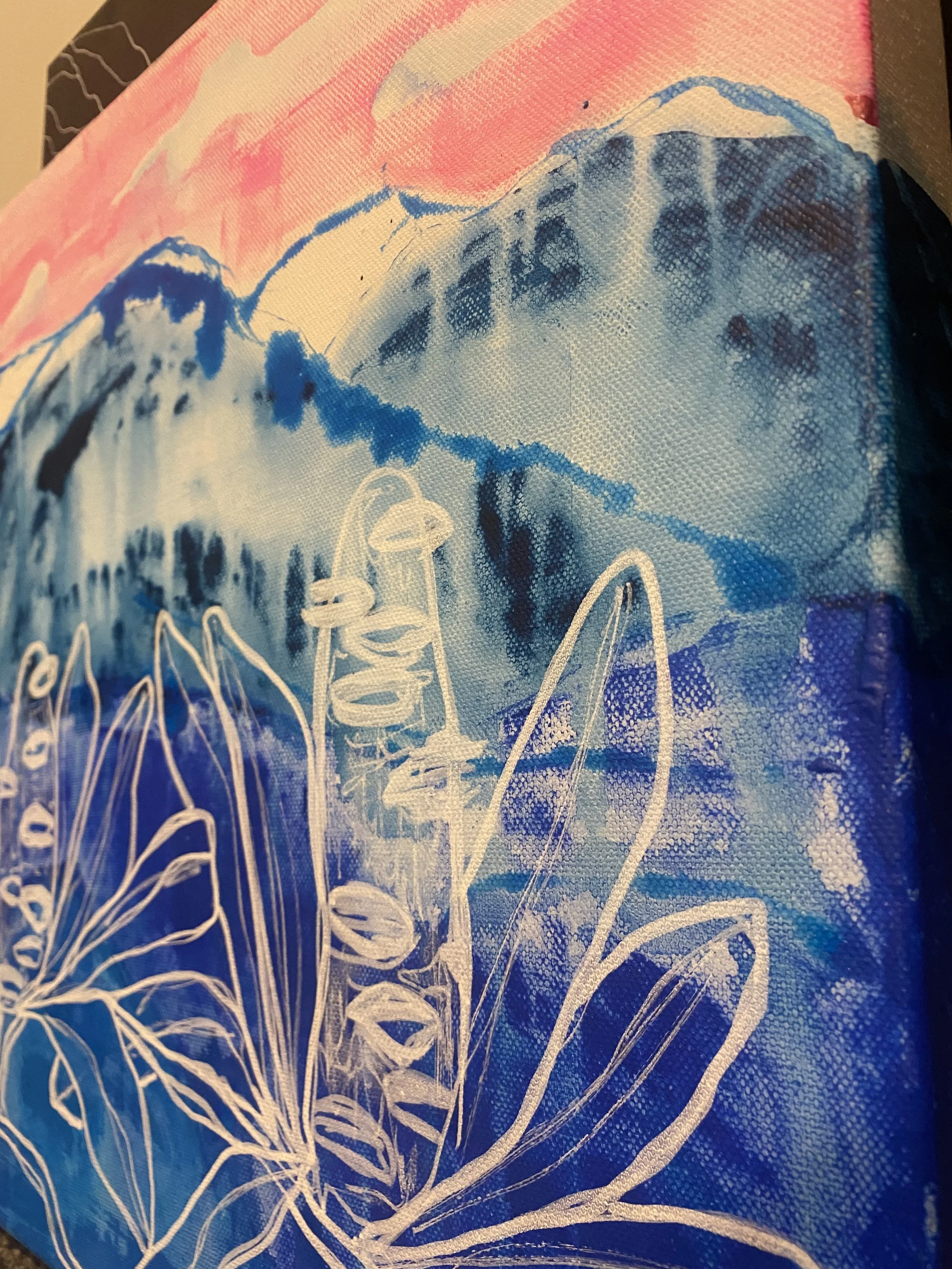 Close-up image of a canvas with an abstract mountain landscape painting in pink, blue, and white hues, featuring a white outline of leaves and plants in the foreground.