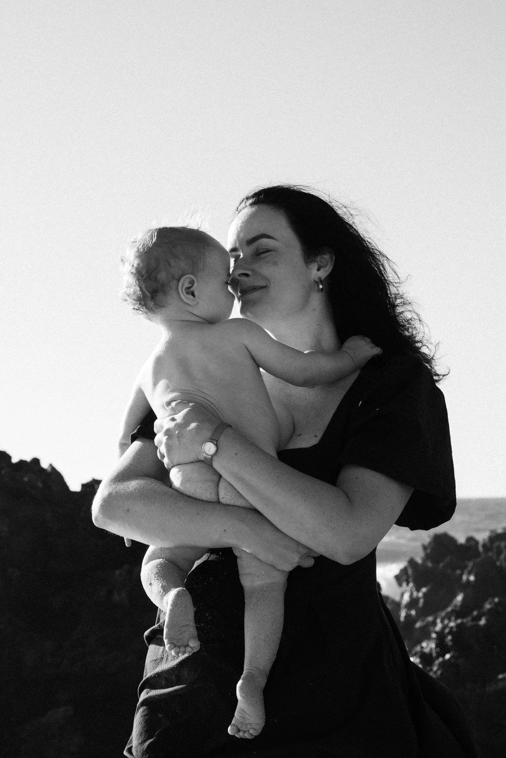 Mother holding her baby close outdoors in Perth, black and white motherhood photography with ocean backdrop