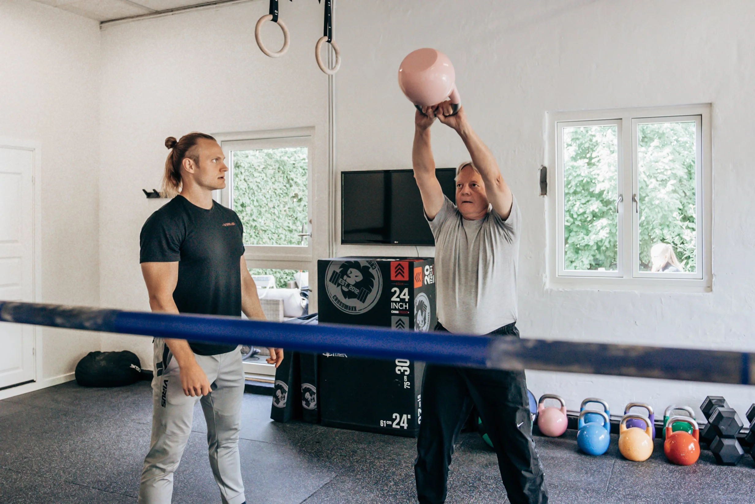 An older man is lifting a pink kettlebell overhead during a workout, while a younger man watches attentively in a gym. The gym has windows, colorful kettlebells, and boxing equipment.