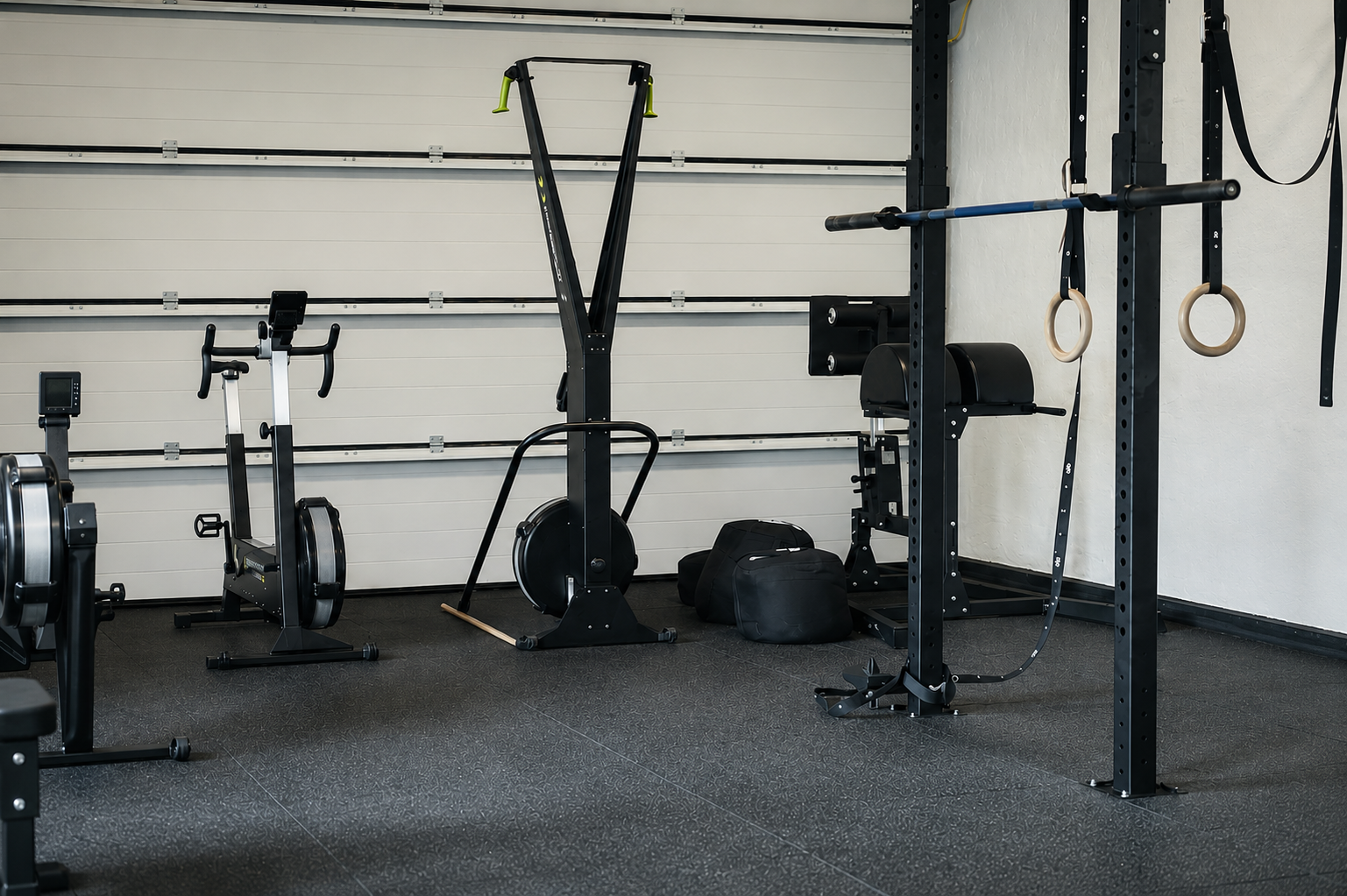 Home gym with exercise bike, rowing machine, gymnastic rings, and weight rack on black rubber flooring.