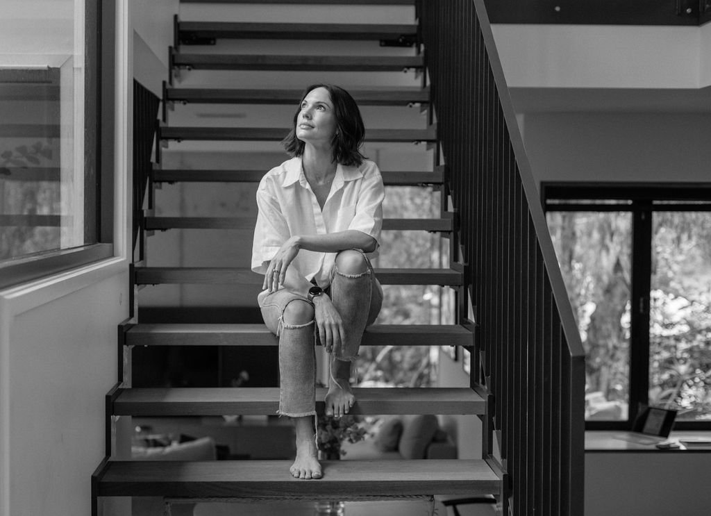A woman sitting barefoot on stairs inside a modern house, looking upwards with a thoughtful expression.